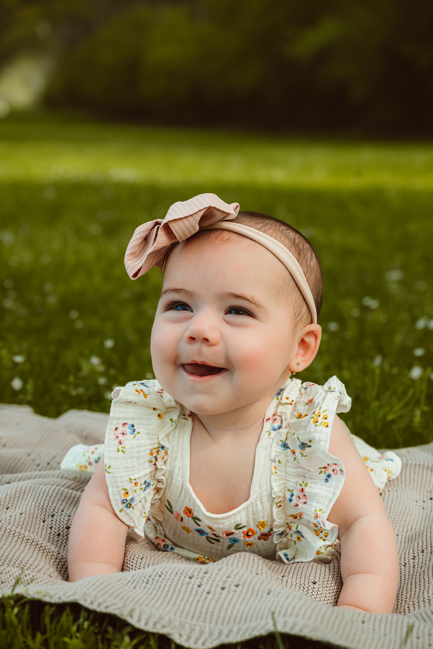 A baby wearing a floral outfit and pink bow headband smiles while lying on a blanket outdoors.
