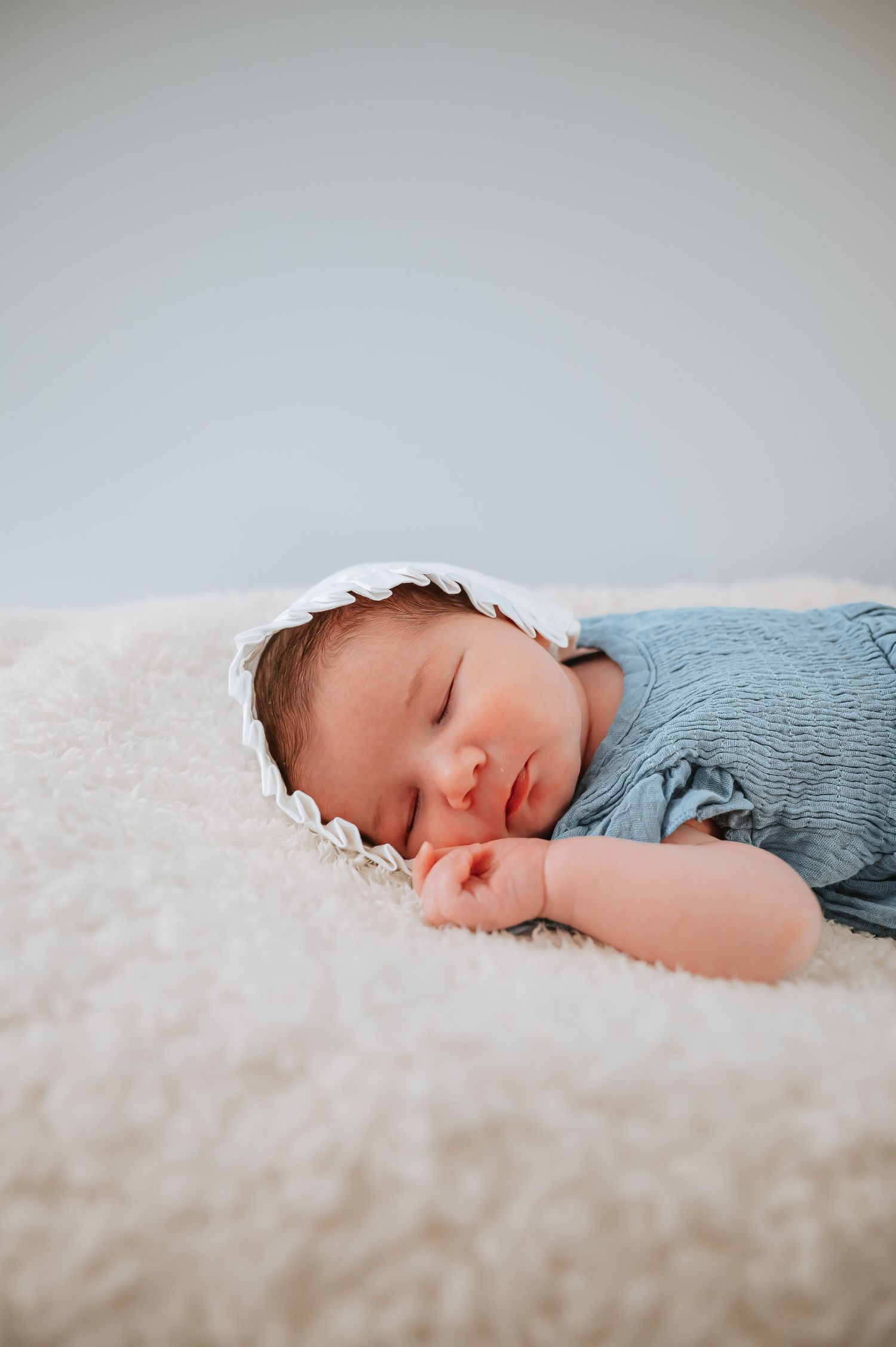 A sleeping newborn baby wearing a blue outfit and white bonnet rests peacefully on soft white fabric.