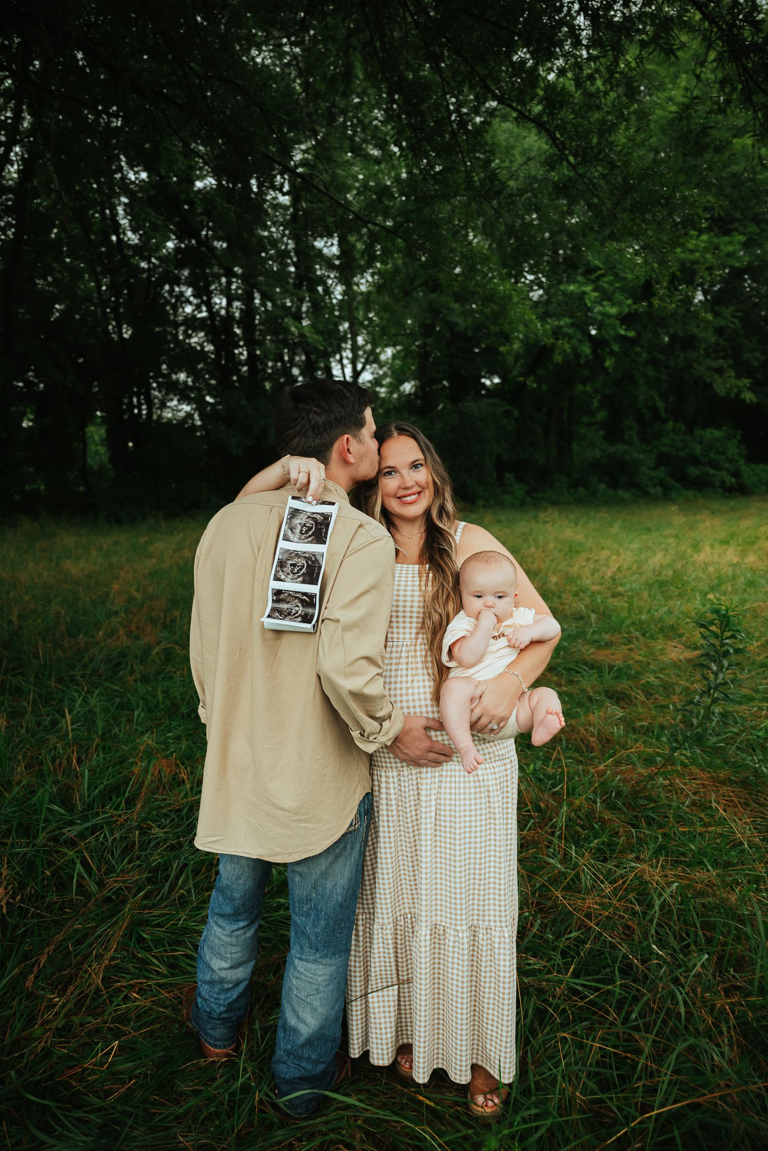 Outdoor family photo session with baby and ultrasound pictures in a green field.