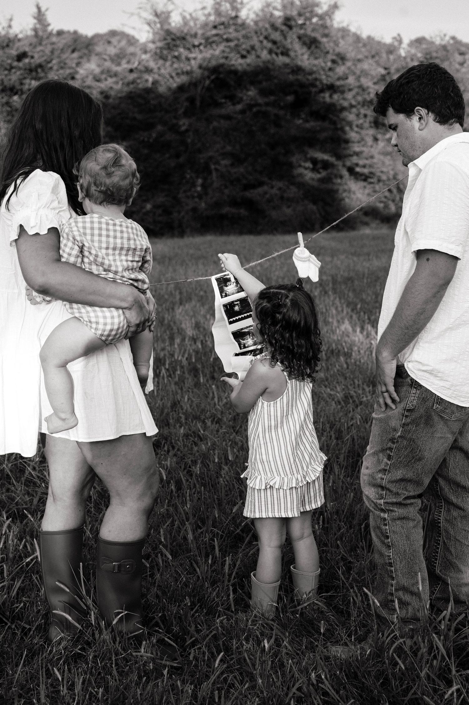Black and white family photo in a field, parents and two children holding ultrasound photos on a string.