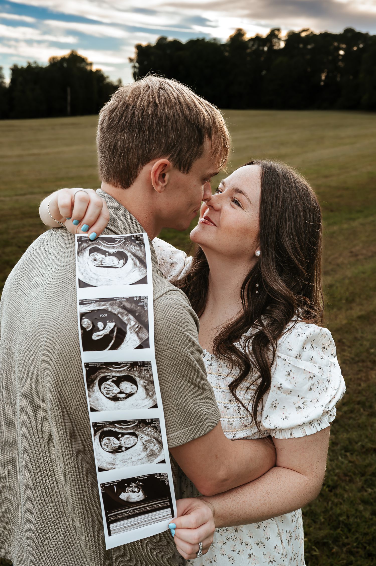 A couple shares an intimate moment while holding ultrasound photos in a field.