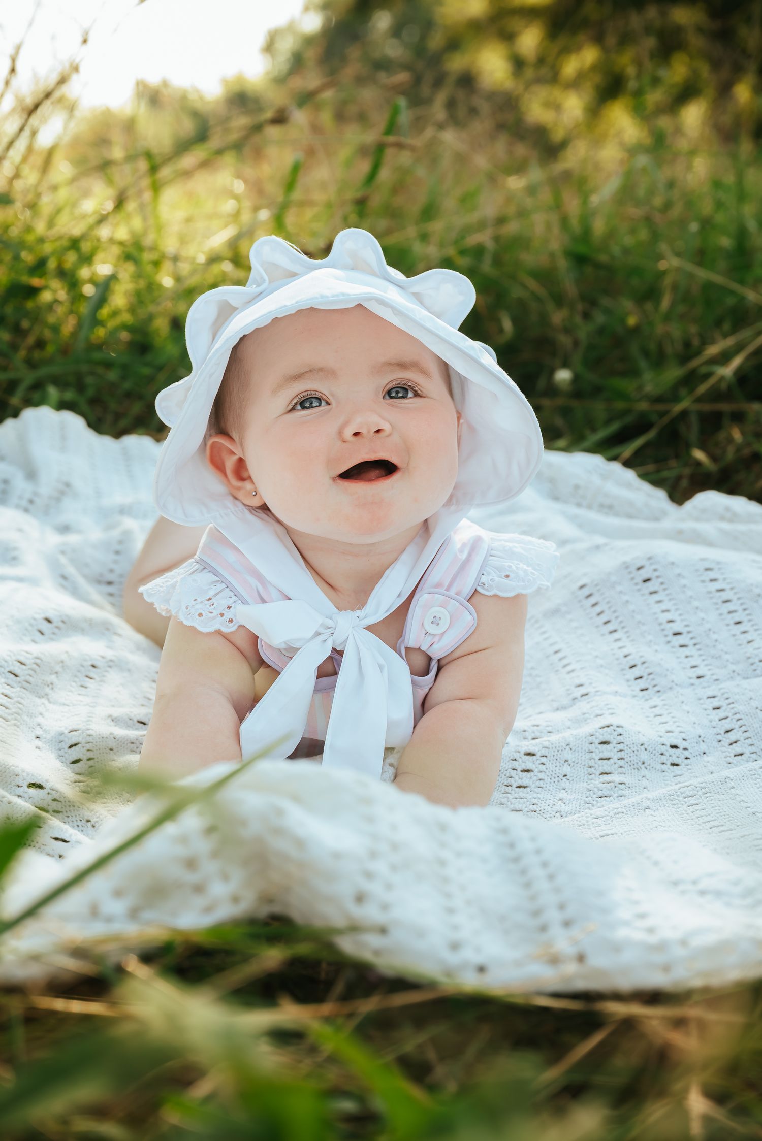 Baby in white bonnet and dress lying on lace blanket in sunlit grass.