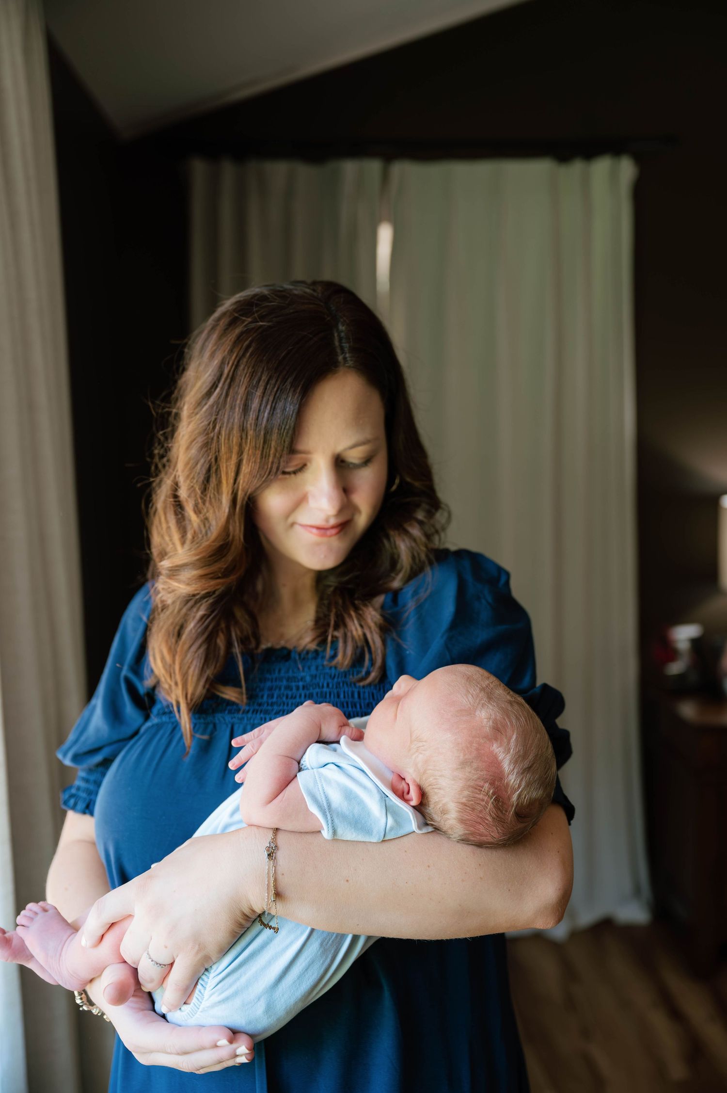 A mother in a blue dress tenderly gazes at her newborn baby cradled in her arms indoors.
