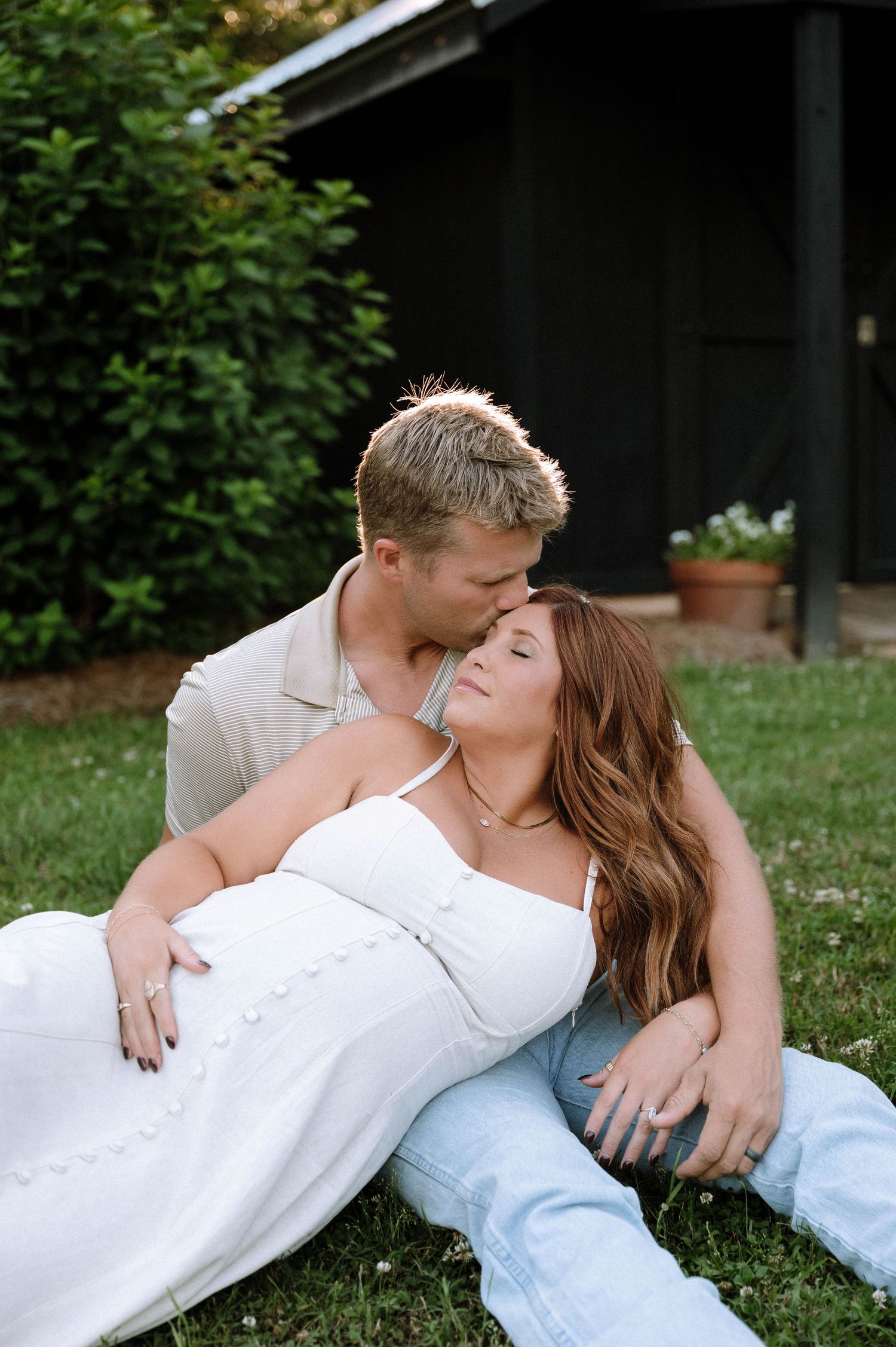 A couple sits on grass, man kissing woman's forehead as she leans back in white dress and jeans.