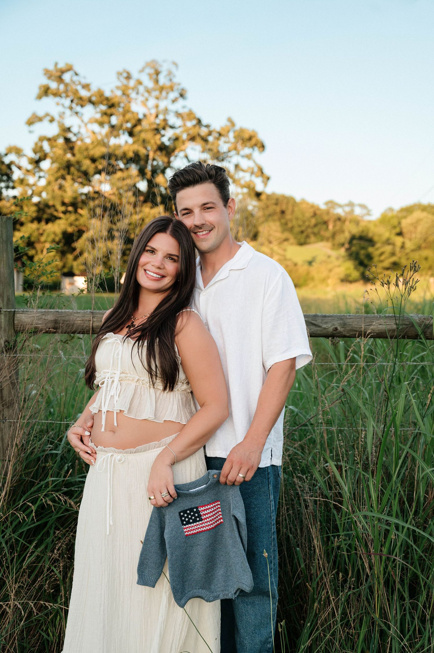 Couple poses outdoors holding American flag baby onesie, announcing pregnancy near rustic fence and green trees.