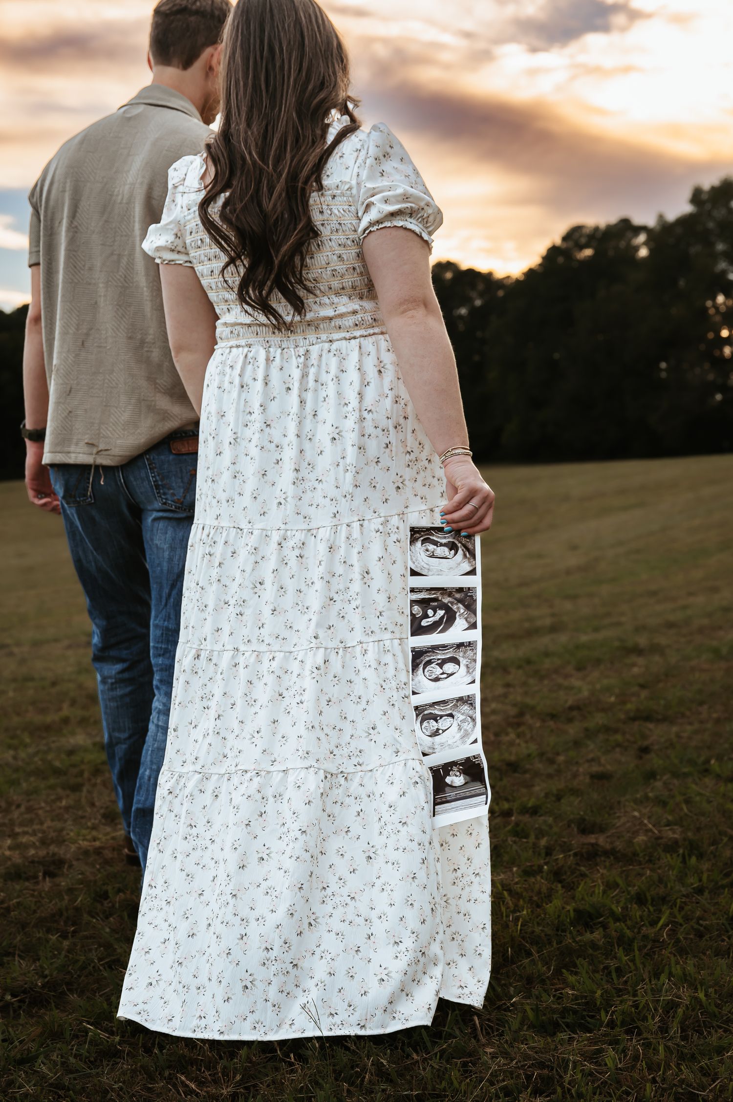 A couple stands in a field at sunset holding ultrasound photos.