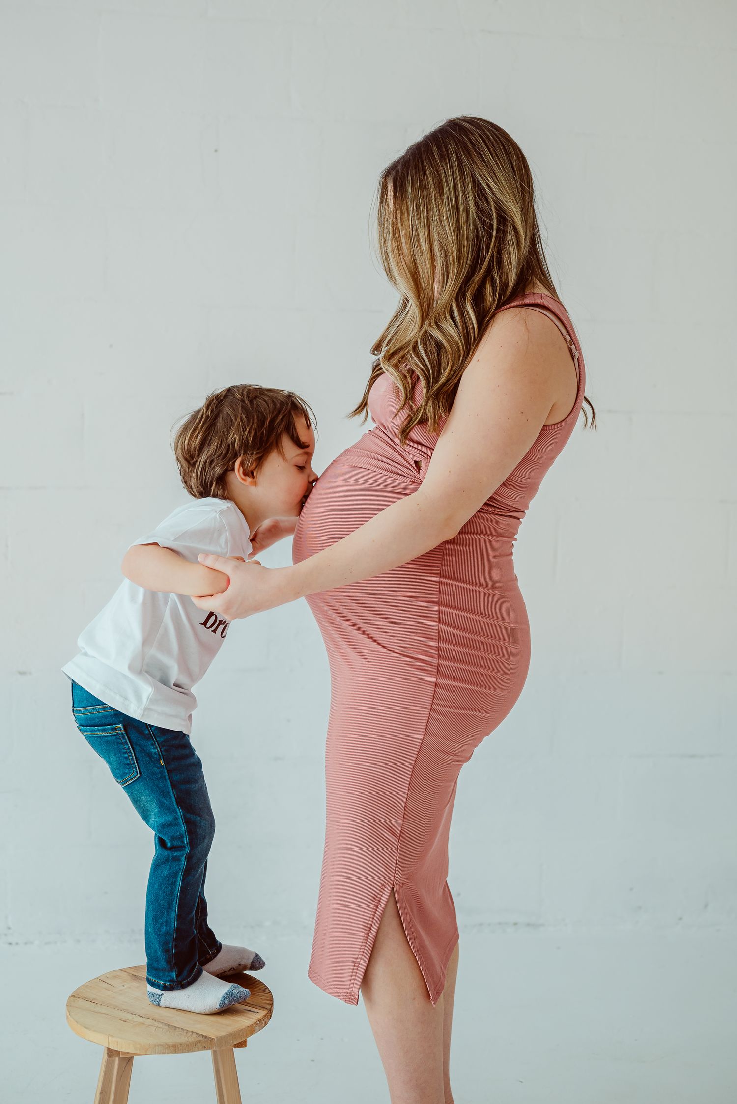 Touching maternity moment with child reaching up to pregnant belly in pink dress.