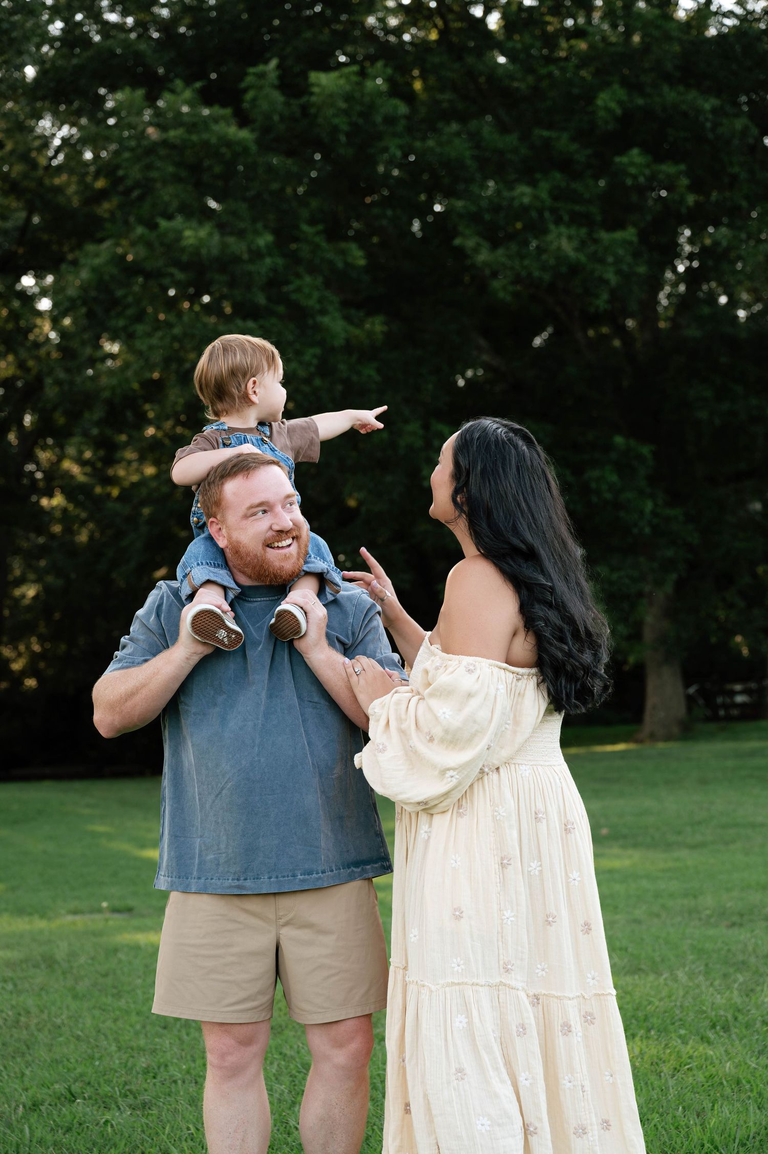 A happy family with a toddler on dad's shoulders in a lush green park, mom in a flowy yellow dress smiling.