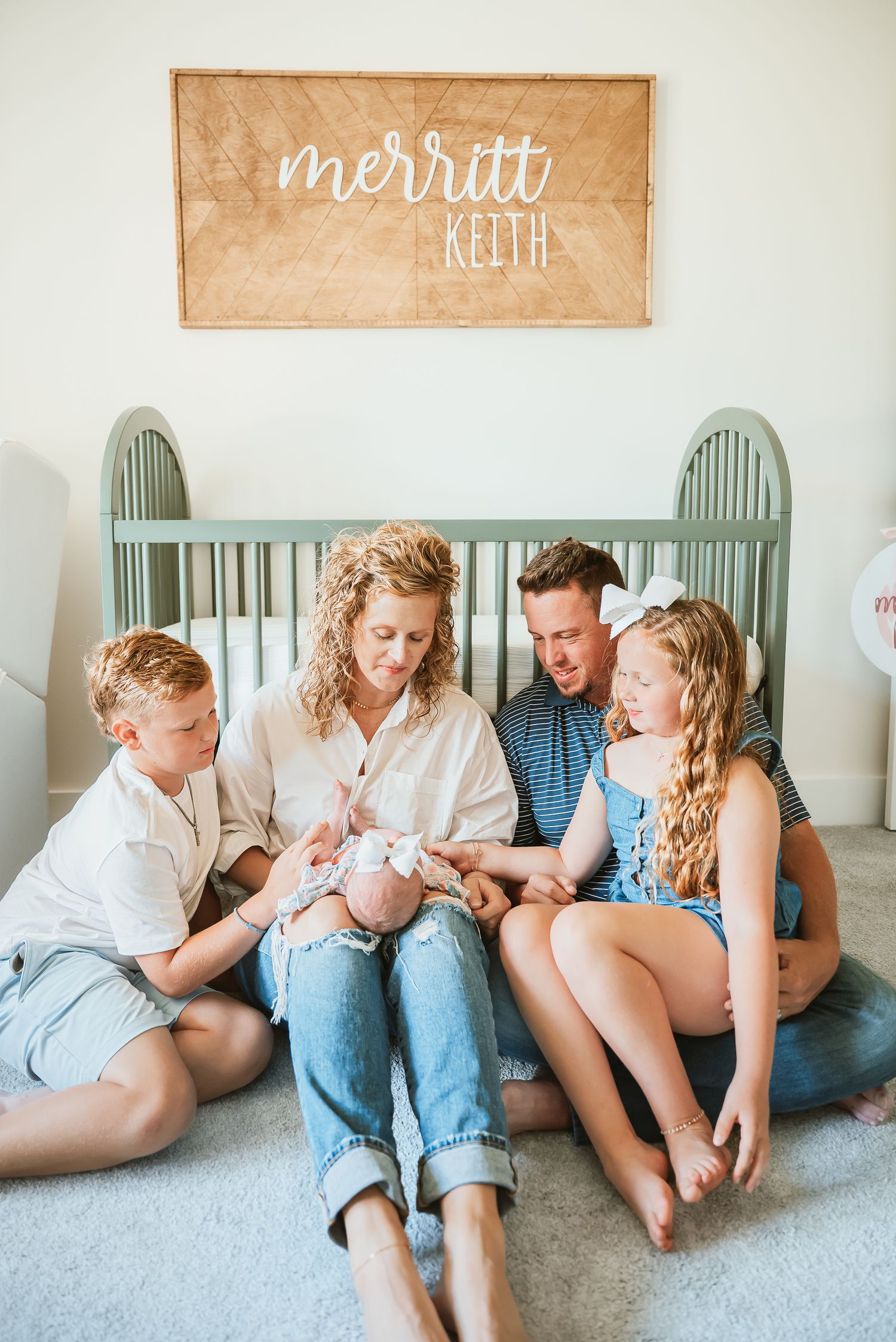 A family sits together on the floor in front of a mint green crib with a wooden name sign above.