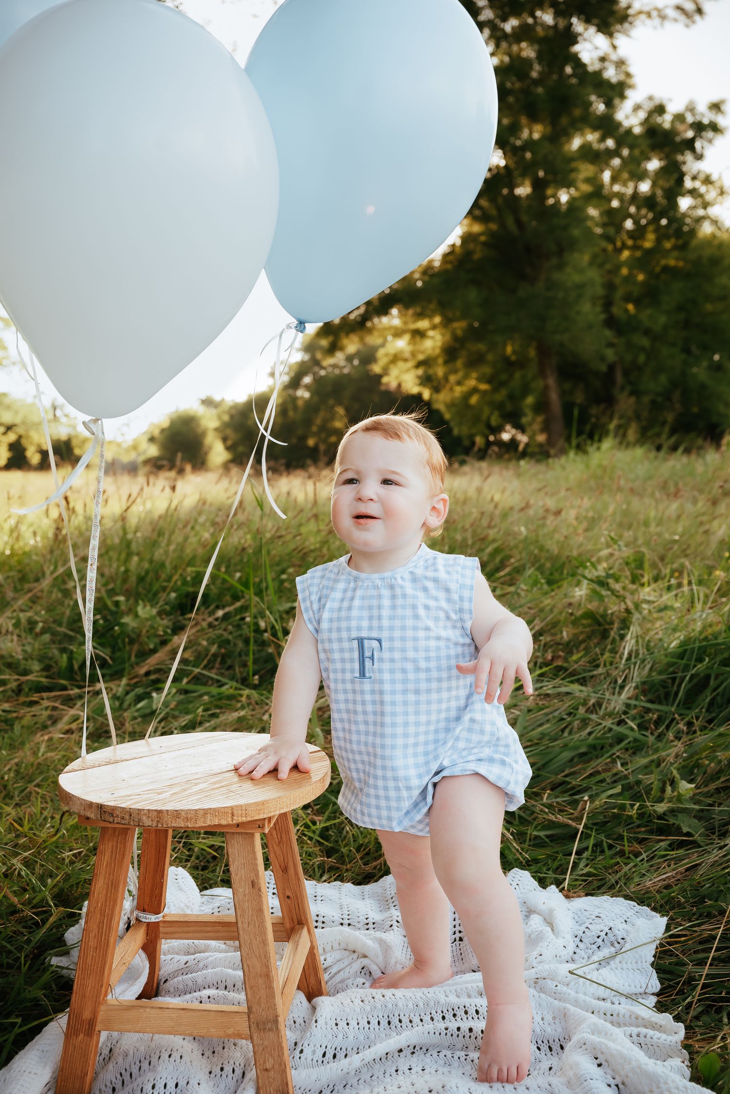 Child in blue gingham outfit standing on wooden stool with balloons in field.