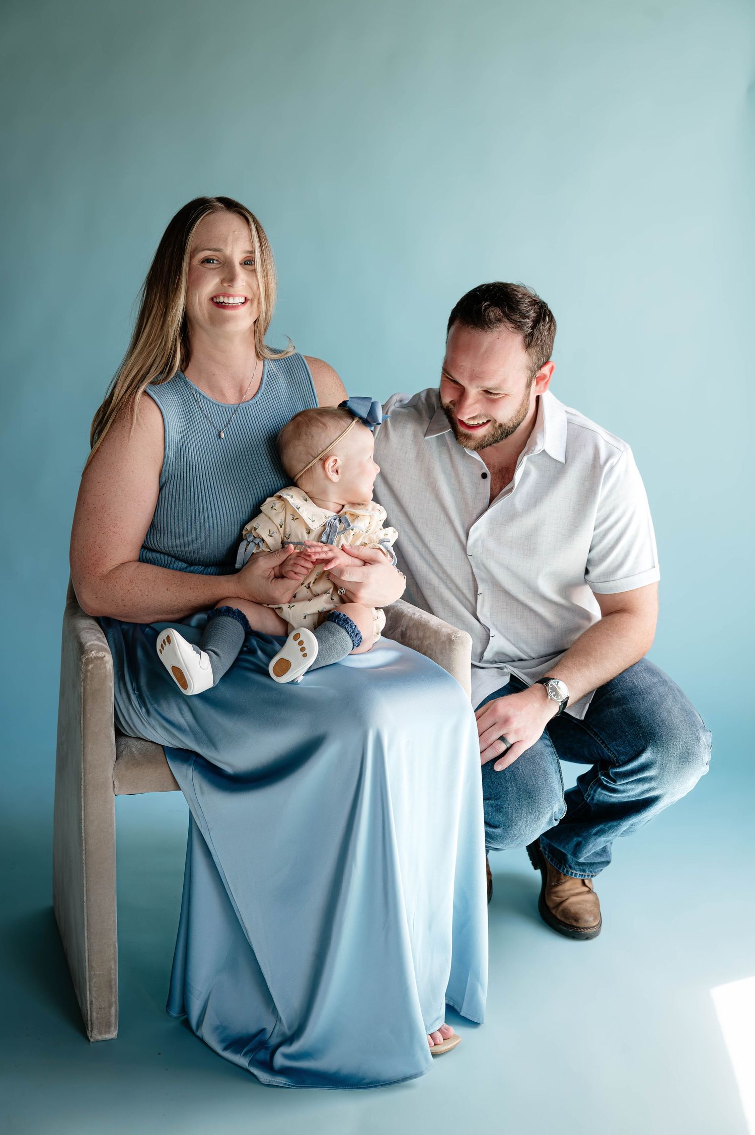 Happy family portrait with mother in blue dress holding baby while father kneels beside them on blue background.