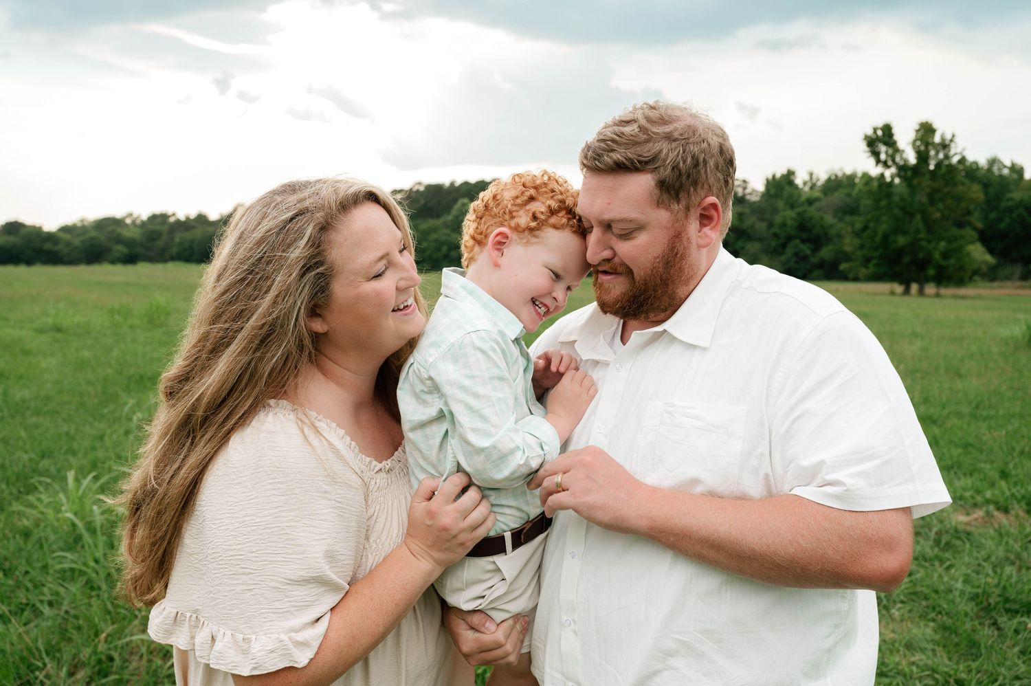 A joyful family of three poses in a green field, parents smiling at their red-haired toddler son on a cloudy day.