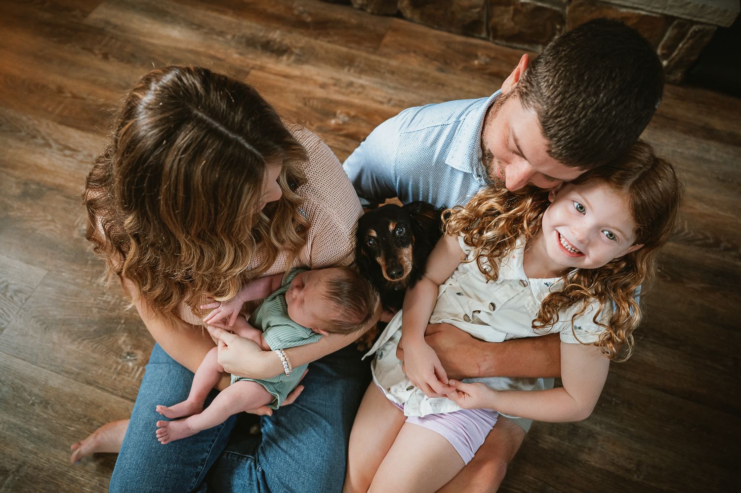 A family cuddles together on a wooden floor while holding a small black cat.