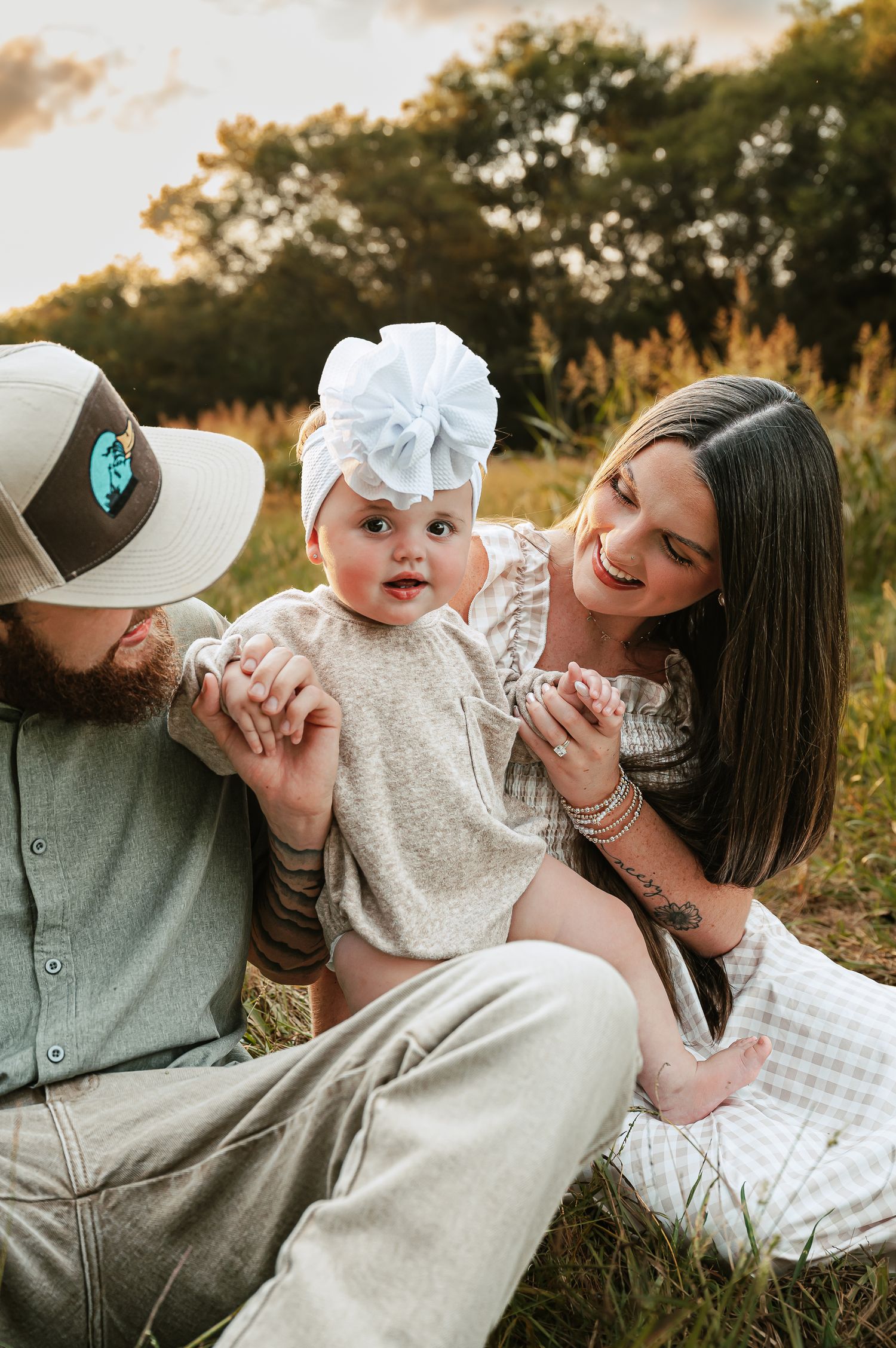 Family with baby in white dress enjoying outdoor photo session at sunset.