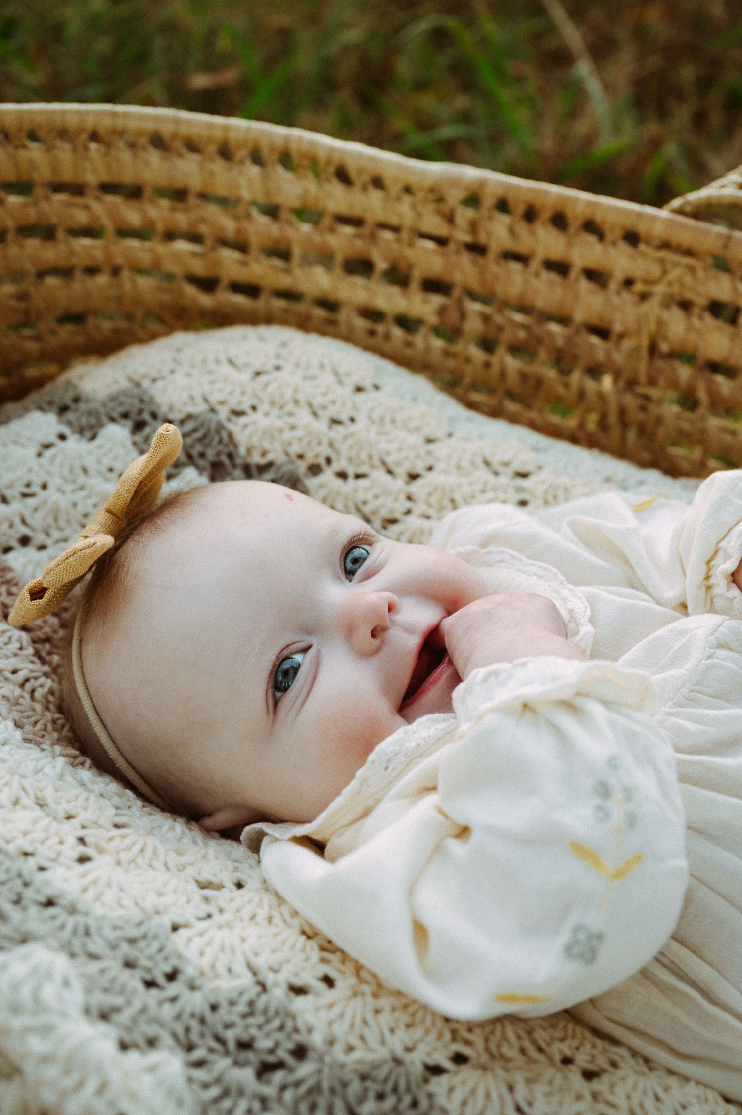 Baby lying in a woven basket on lace fabric wearing a white dress.