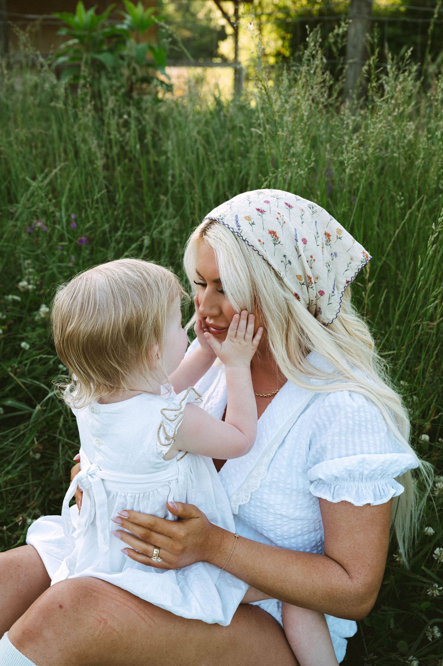 Blonde woman in white dress and floral headscarf holds a toddler girl in white dress outdoors in green field.
