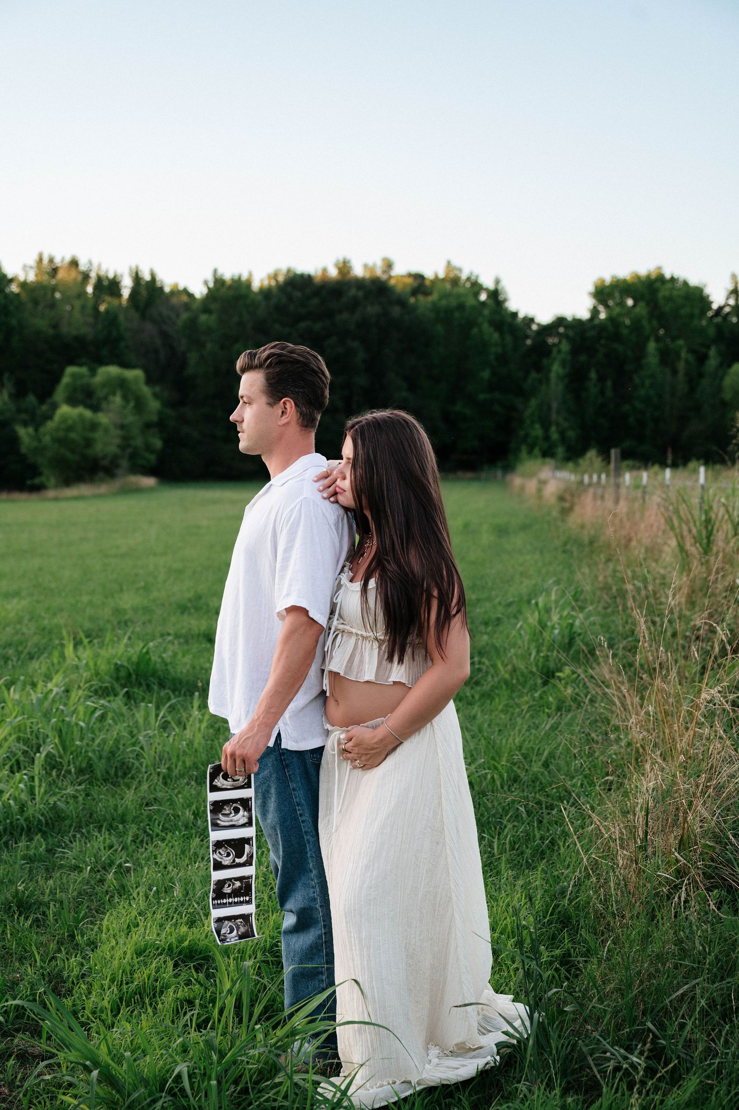Couple standing in green field holding ultrasound photos, woman in white maxi dress embracing man in white shirt and jeans.
