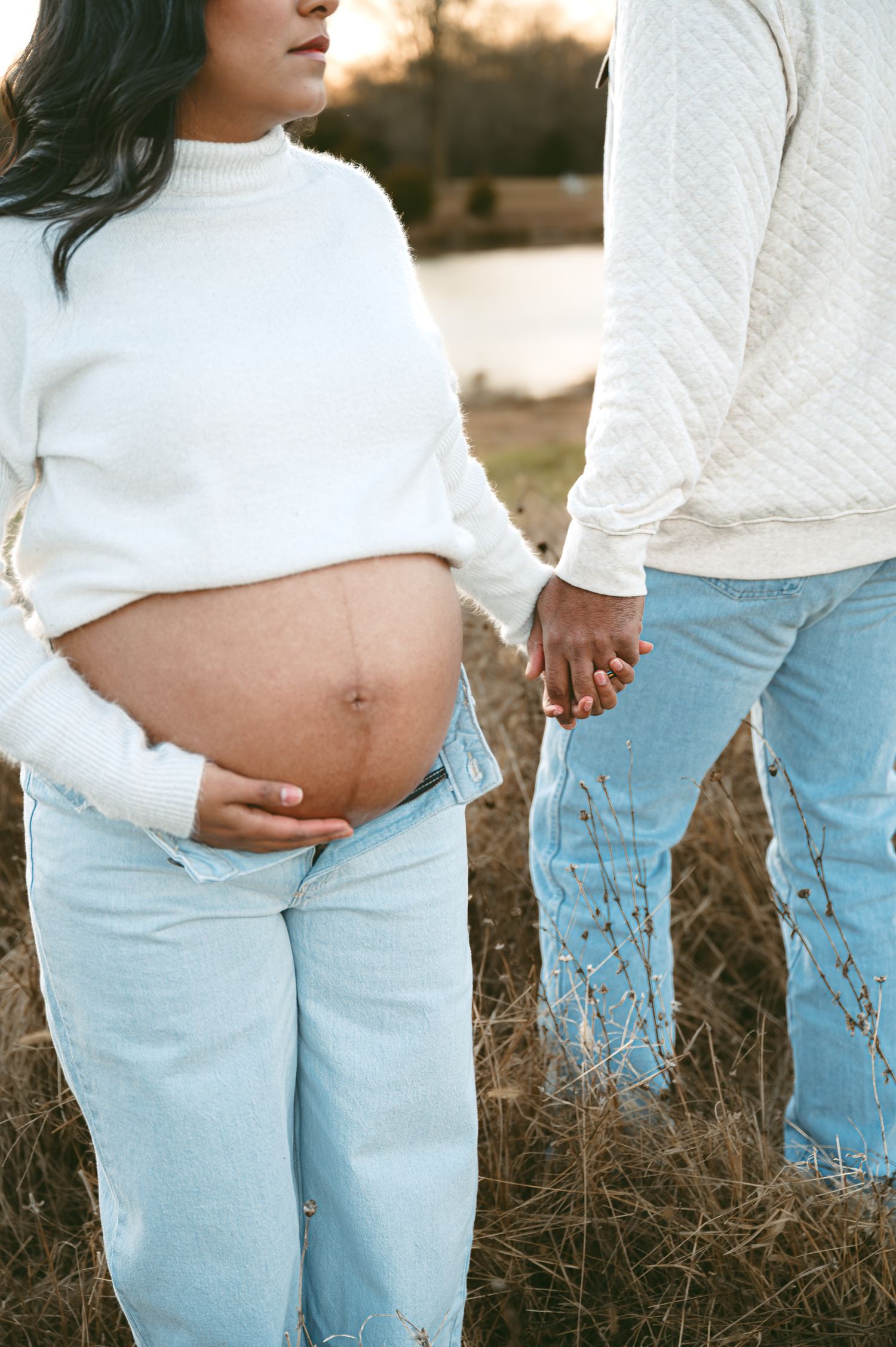 Pregnant woman in white sweater and jeans holds hands with partner outdoors in a maternity photo session.