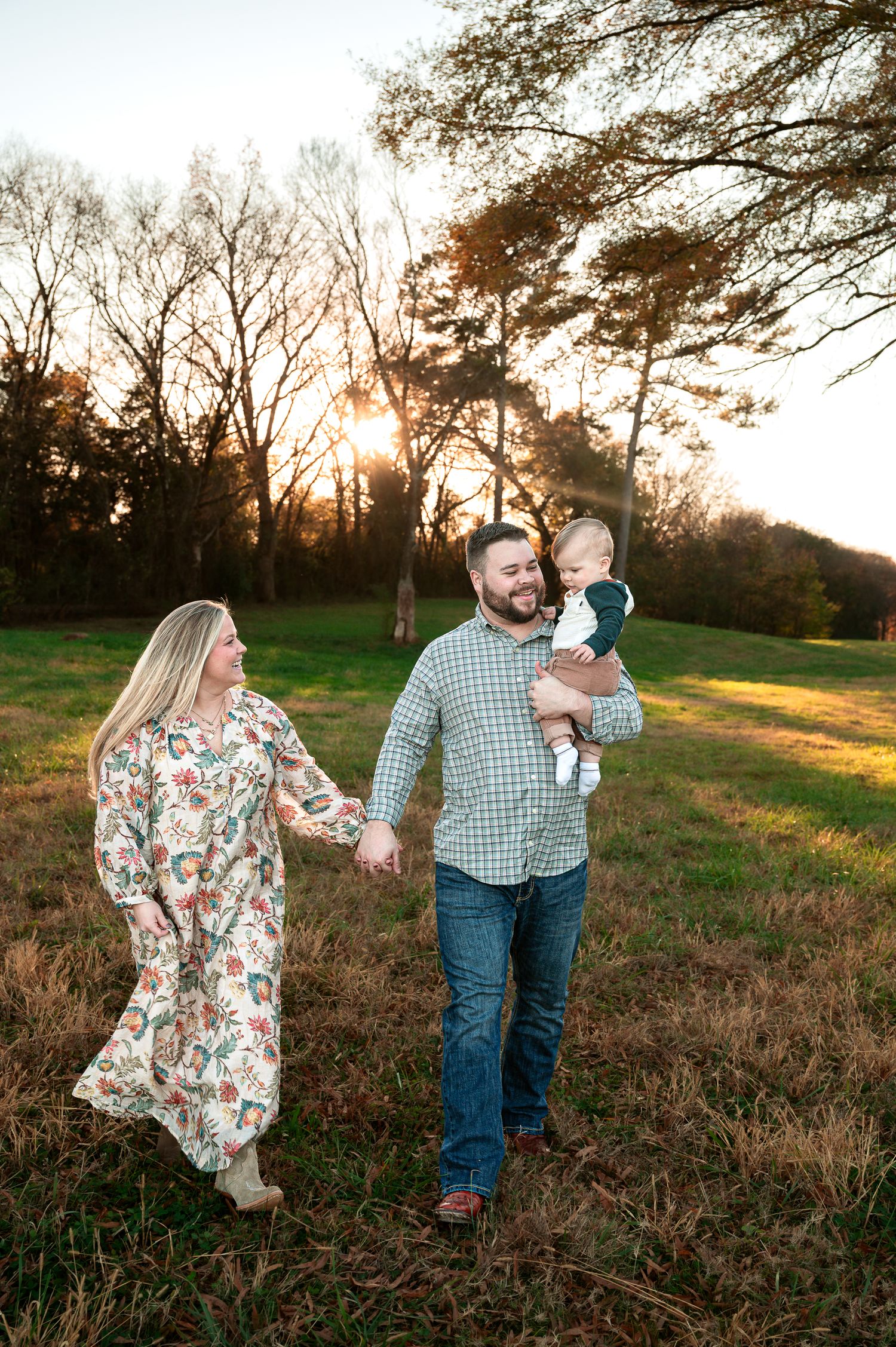 A family walks together at sunset in a grassy field with tall trees in the background.