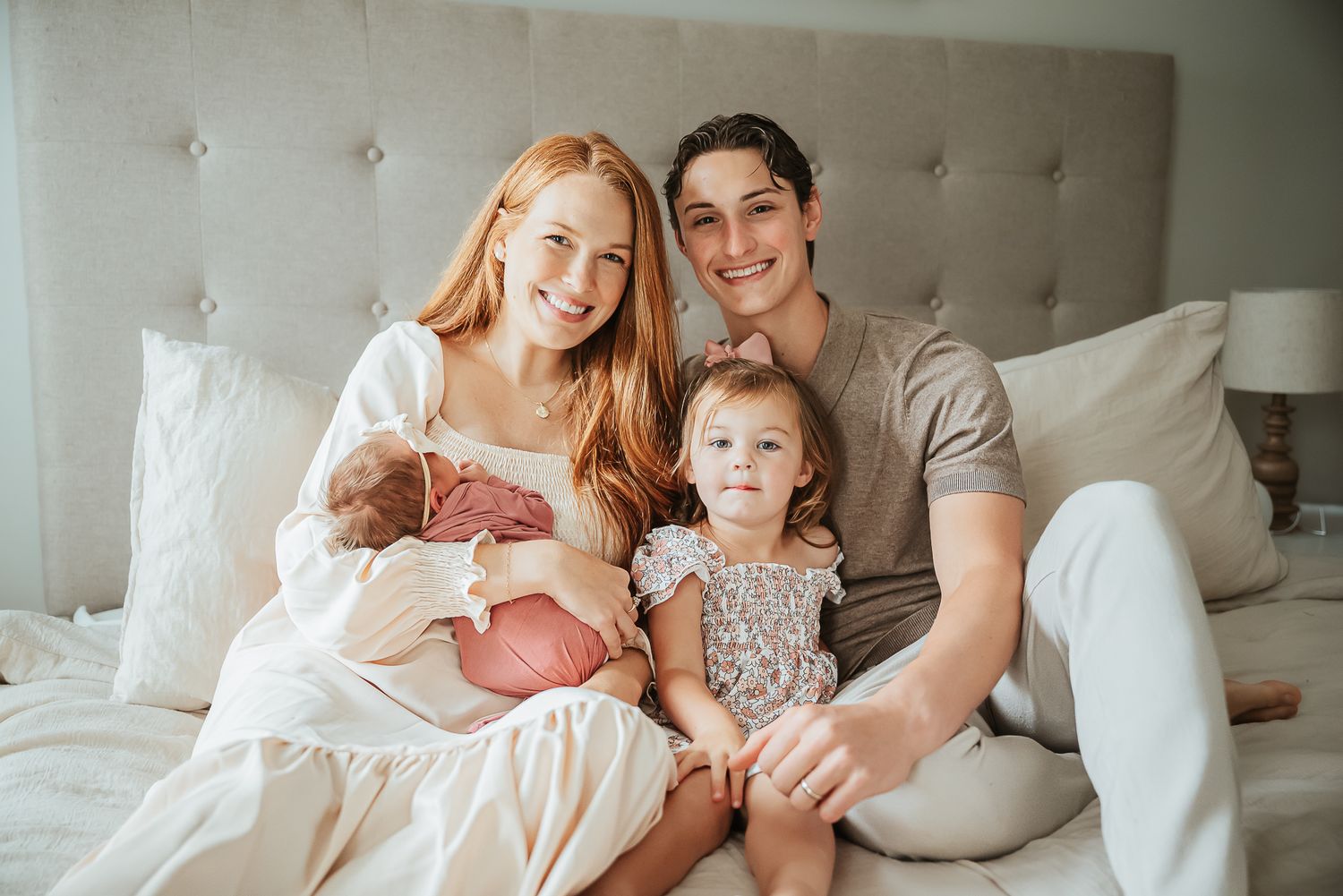 A family sits together on a tufted headboard bed with their newborn baby.