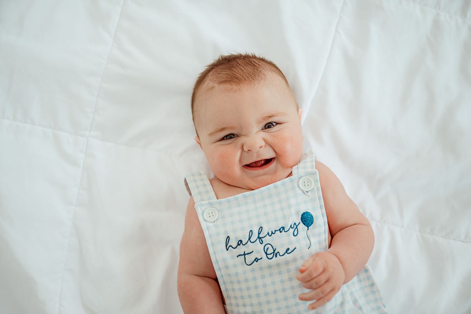 A smiling baby lies on white bedding holding a milestone card.