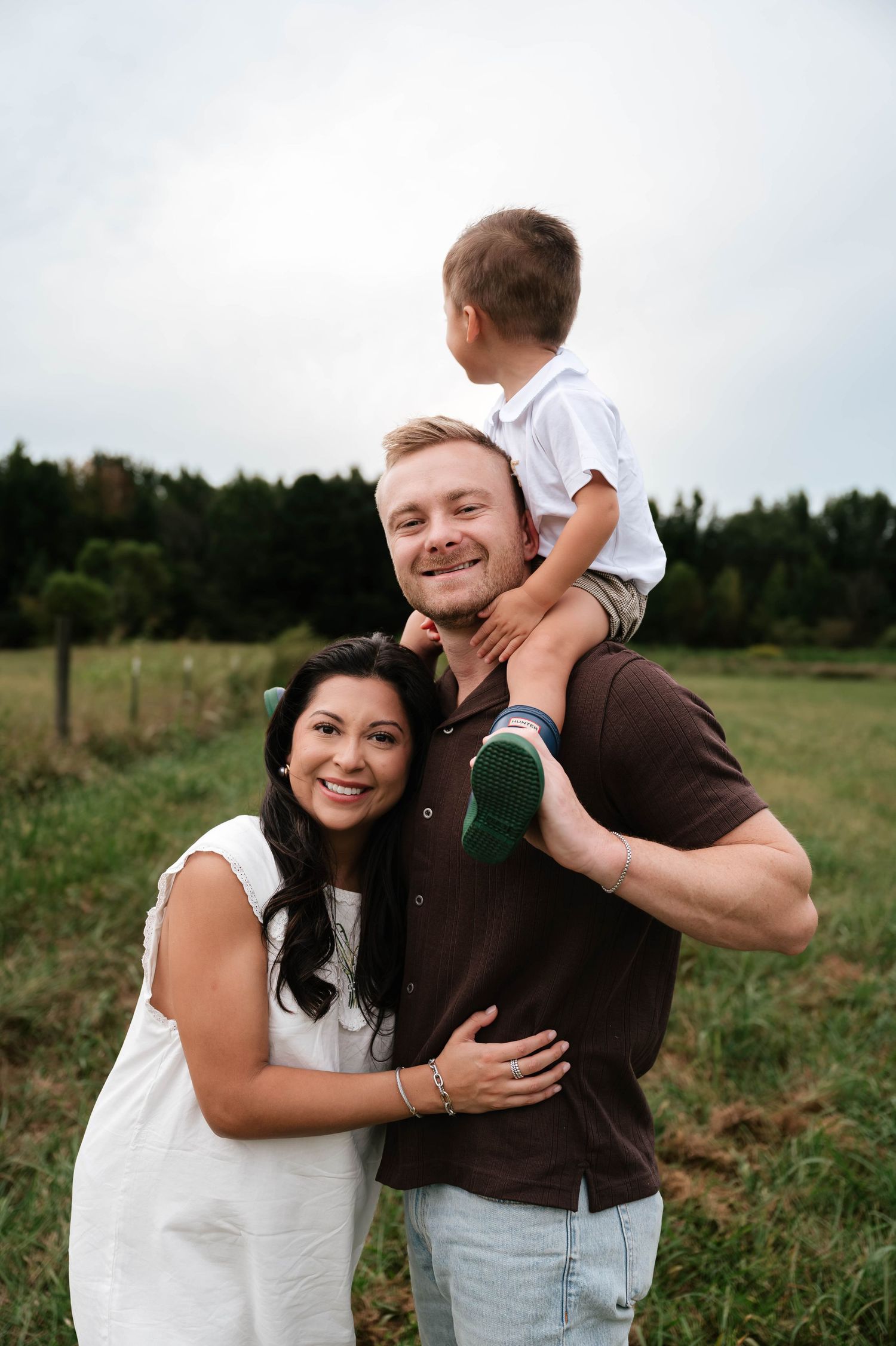A smiling family of three poses outdoors, with a woman in white, a man in brown, and a toddler on his shoulders.