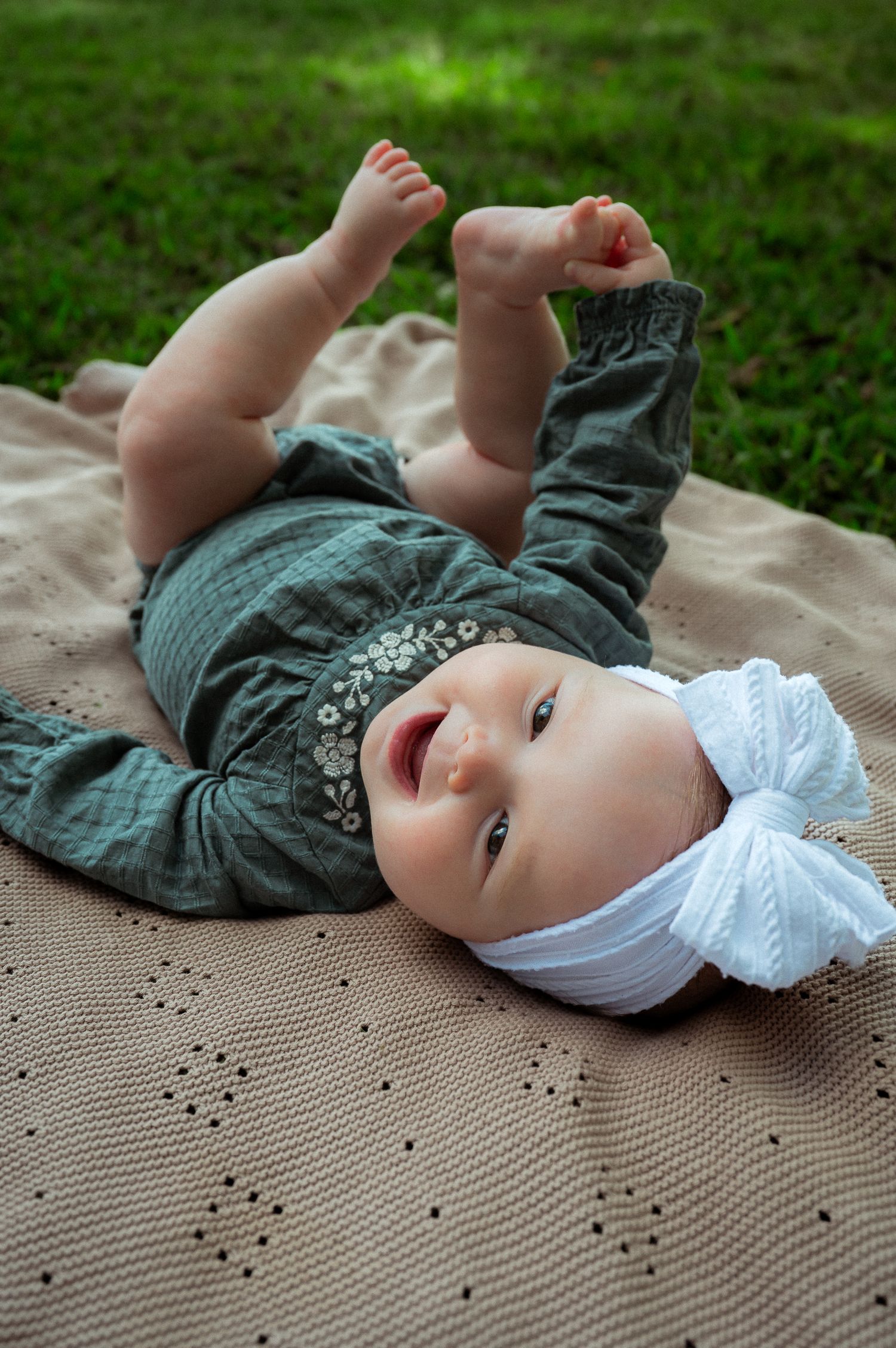 Baby wearing teal outfit and white bow lying on textured blanket outdoors.