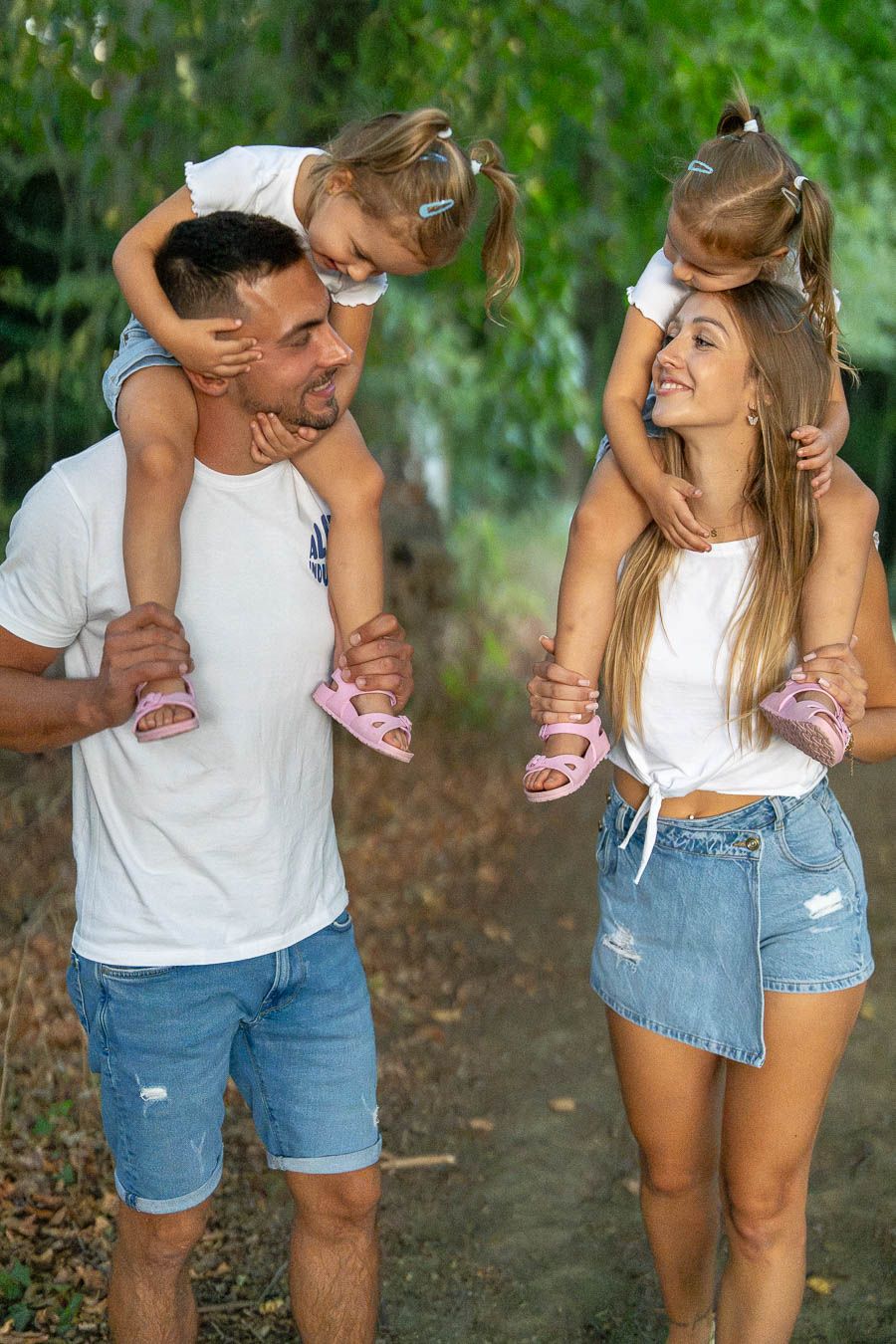 A happy family with two young girls sitting on parents shoulders walking outdoors on a green tree-lined path.