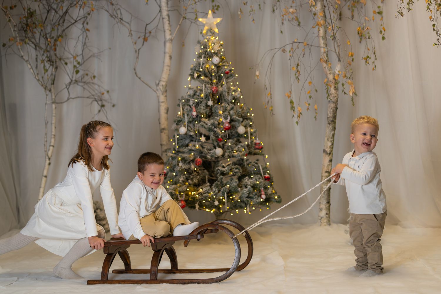 Children play near a vintage wooden sled in front of a decorated Christmas tree.