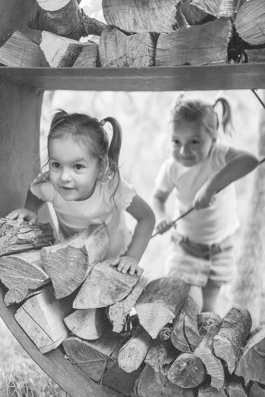 Two young girls with pigtails playing near a rustic firewood stack outdoors in black and white photo.