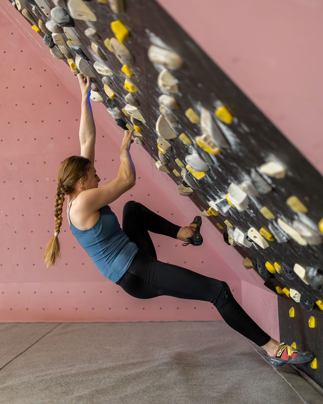 Indoor climber traversing an angled climbing wall with colorful holds against a pink background.