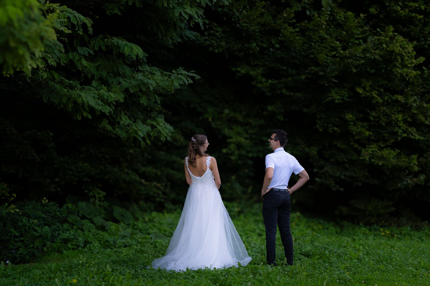 A couple walks through lush greenery wearing wedding attire on a summer day.