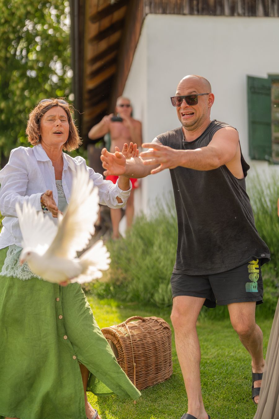 A man and woman release a white dove outdoors on a sunny day near a wicker basket on green grass.
