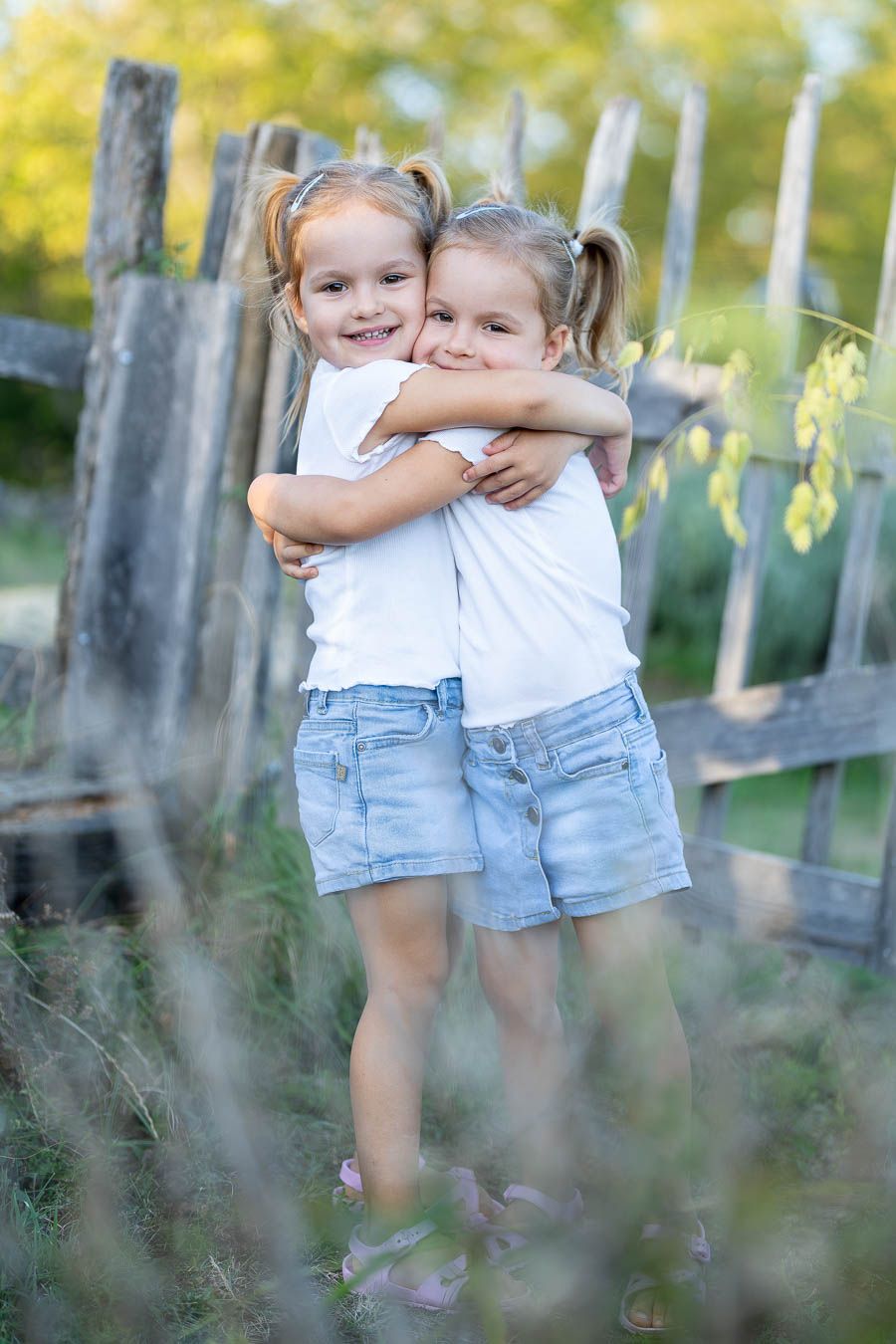 Two young girls in white shirts and denim shorts hugging outdoors near a wooden fence.