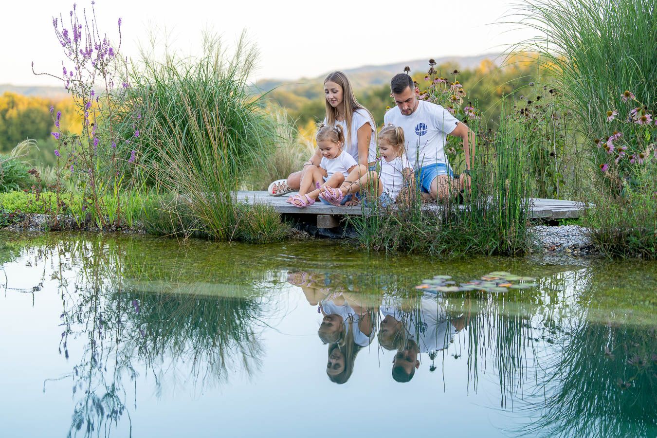 Family of four sitting by a serene reflective pond surrounded by lush greenery, enjoying nature together outdoors.
