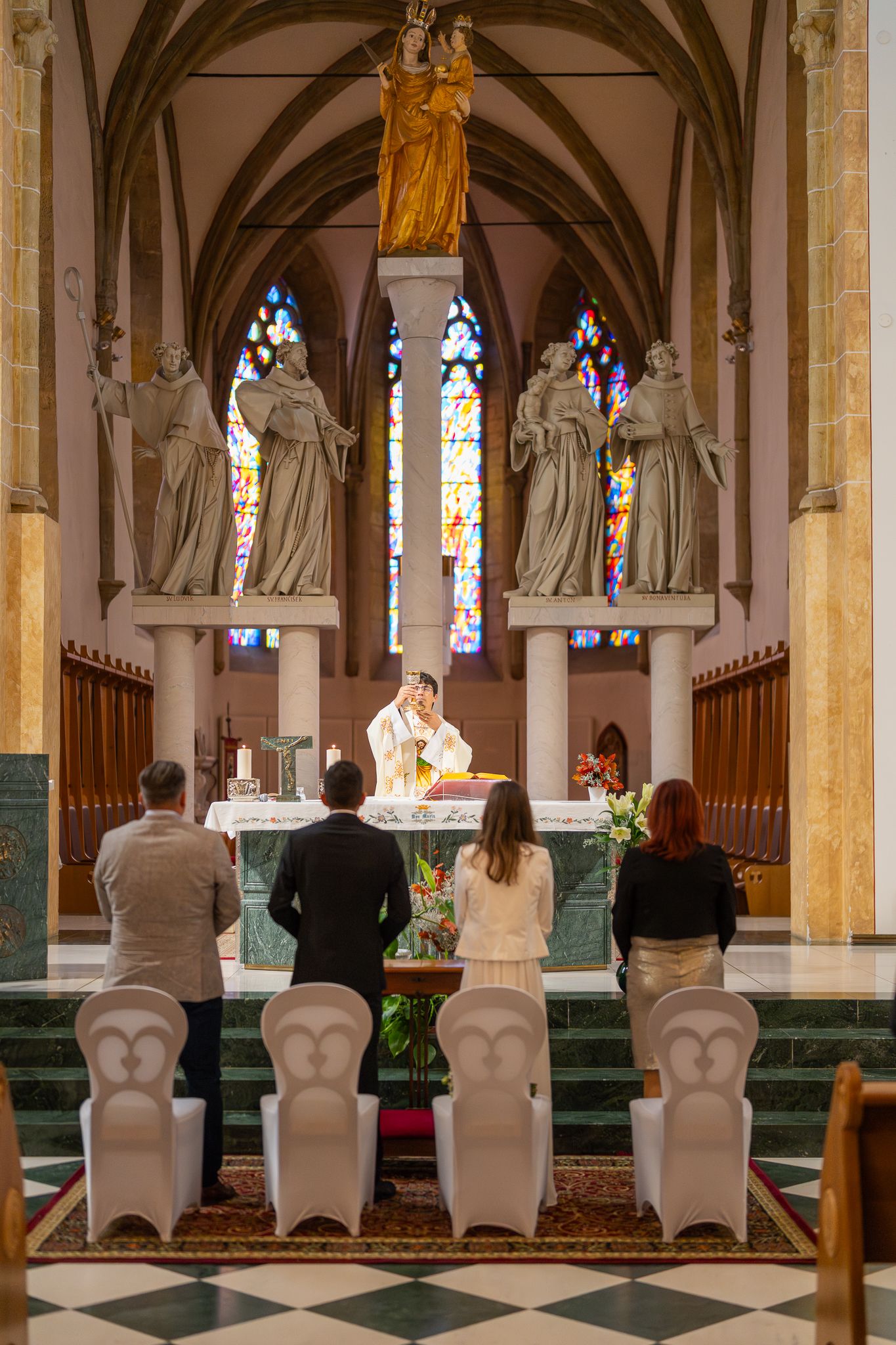 Church ceremony with guests seated in white chairs facing the ornate altar and stained glass windows.