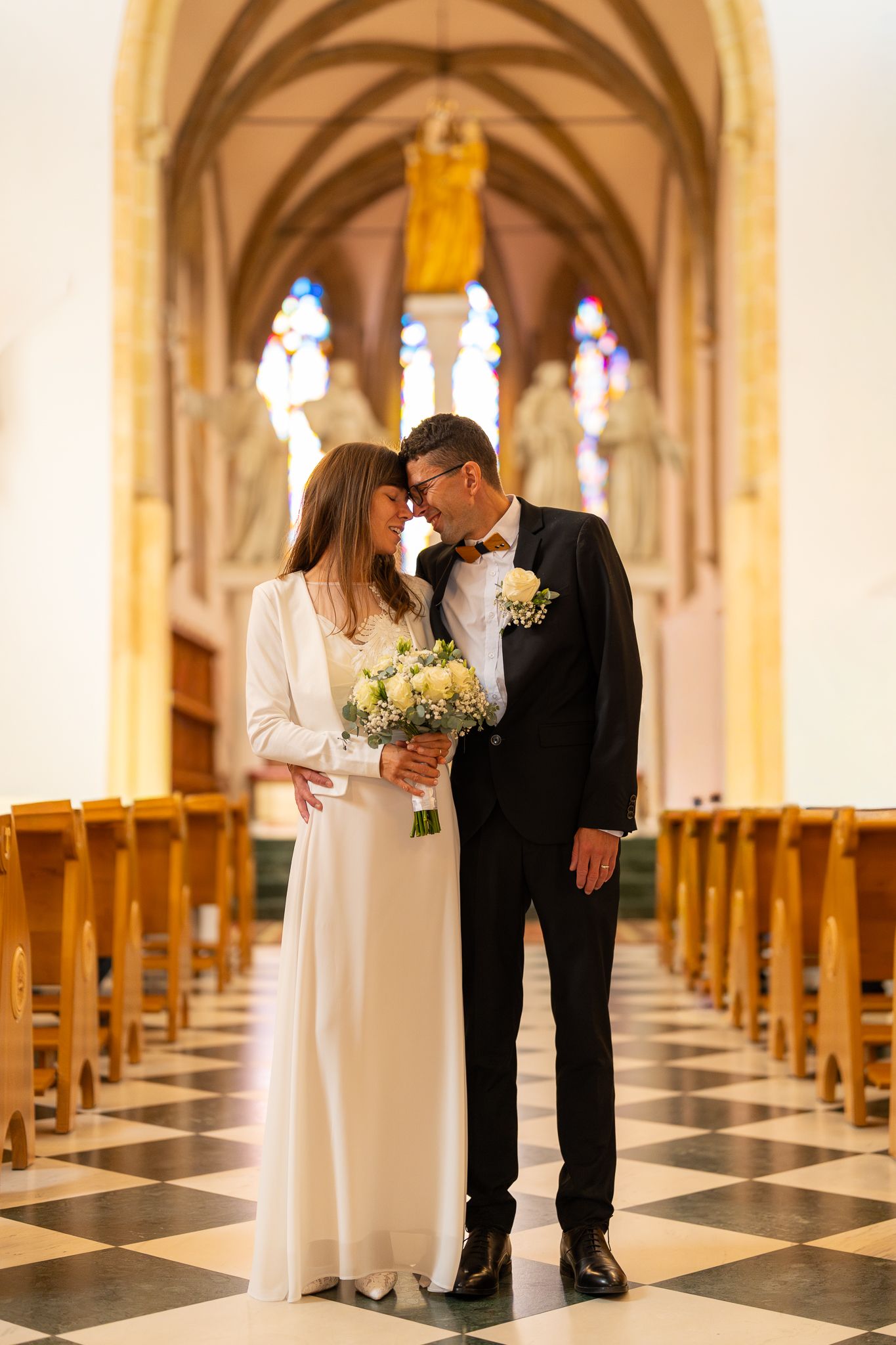 Couple in formal attire poses in church aisle with black and white checkered floor.