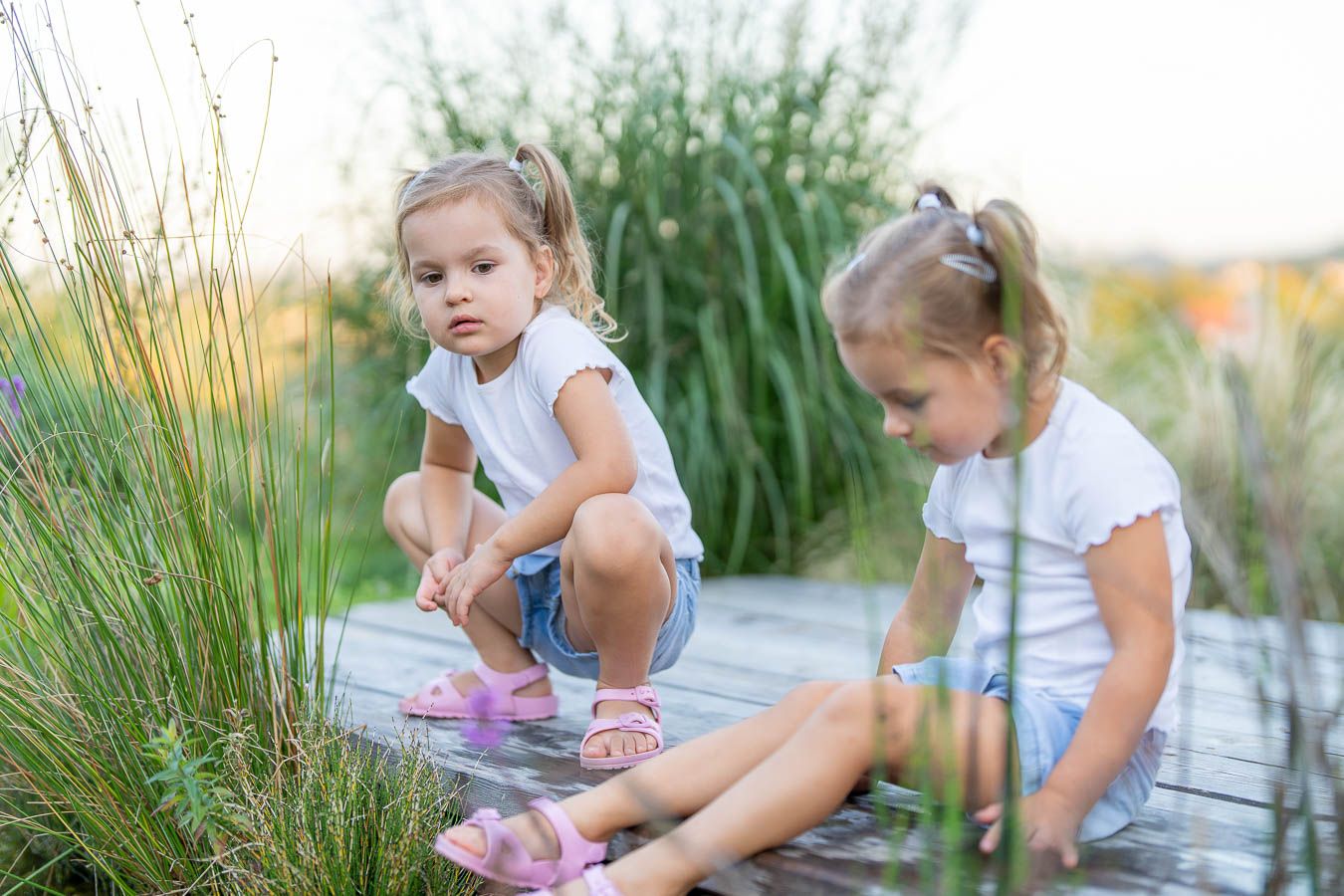 Two little girls in white tops playing on a wooden dock near tall grass and a pond on a sunny day.