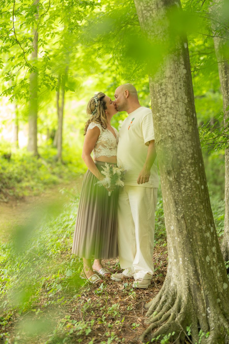 Couple sharing a romantic kiss in a lush green forest, bride holding bouquet in elegant gray skirt and white top.