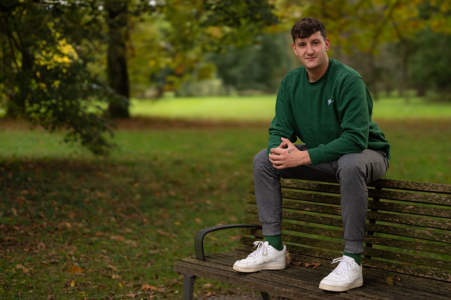Person in green sweatshirt sits casually on park bench surrounded by autumn leaves.