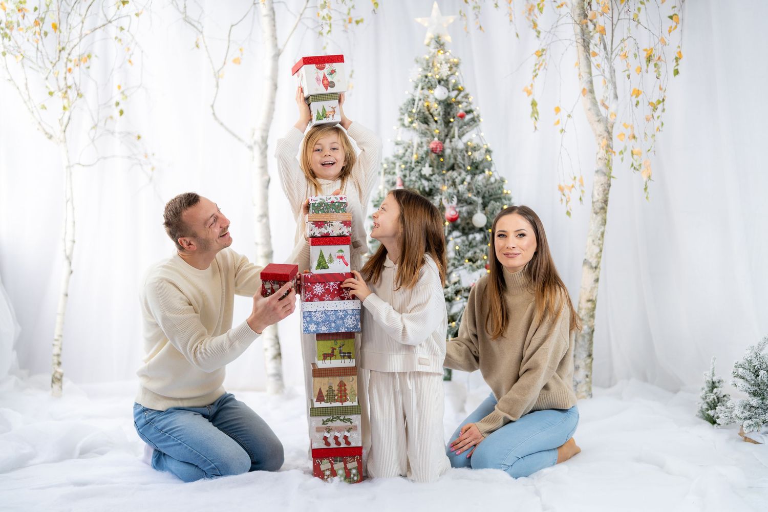 Family poses with tall stack of Christmas presents in white winter wonderland setting with decorated tree.