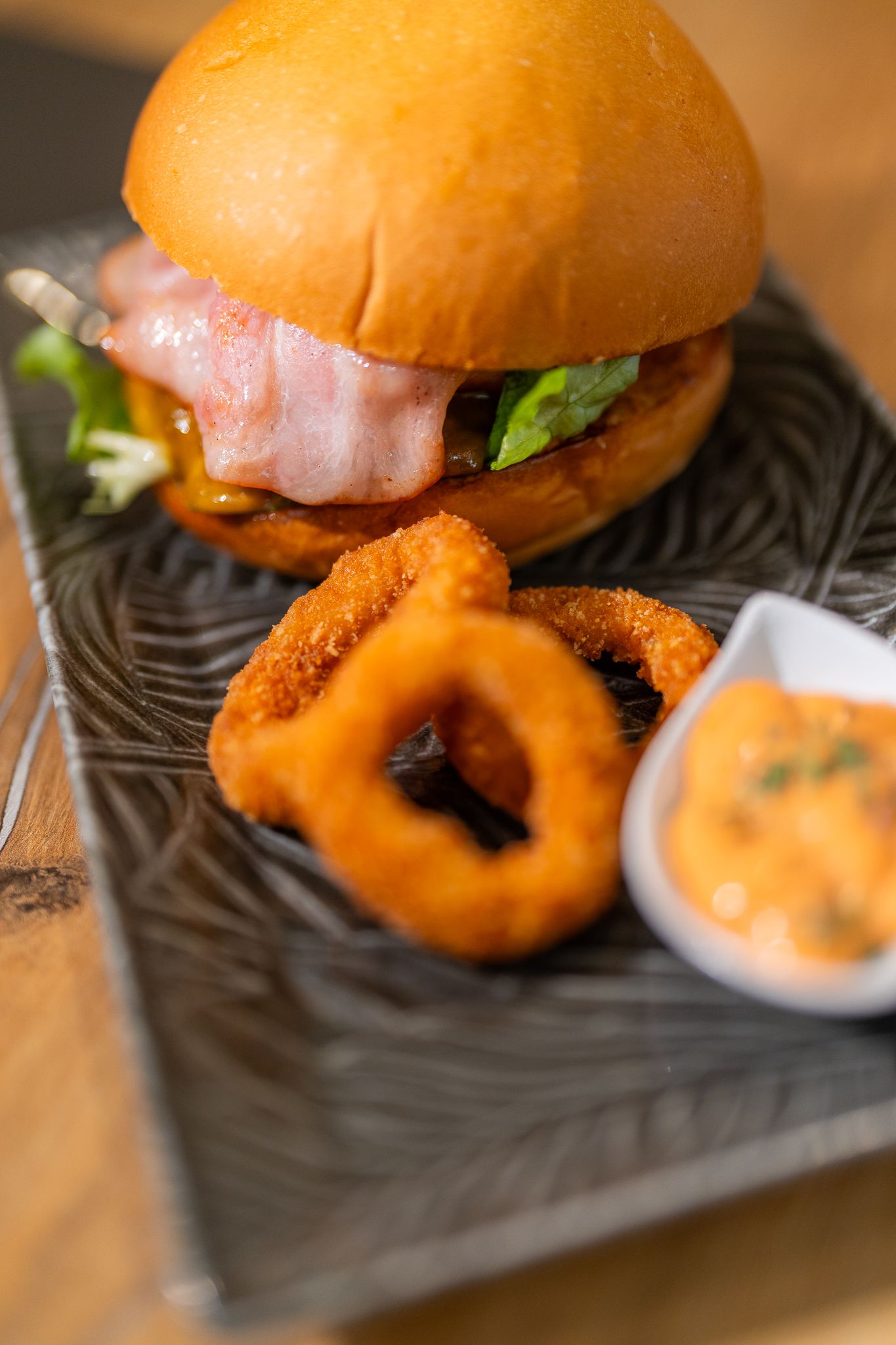 Gourmet burger with onion rings and spicy sauce served on black slate plate.