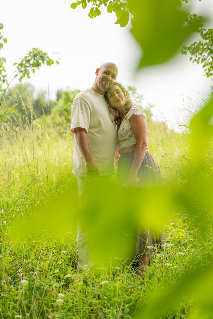 A couple embracing in a lush green meadow, framed by leaves, smiling warmly on a bright summer day.
