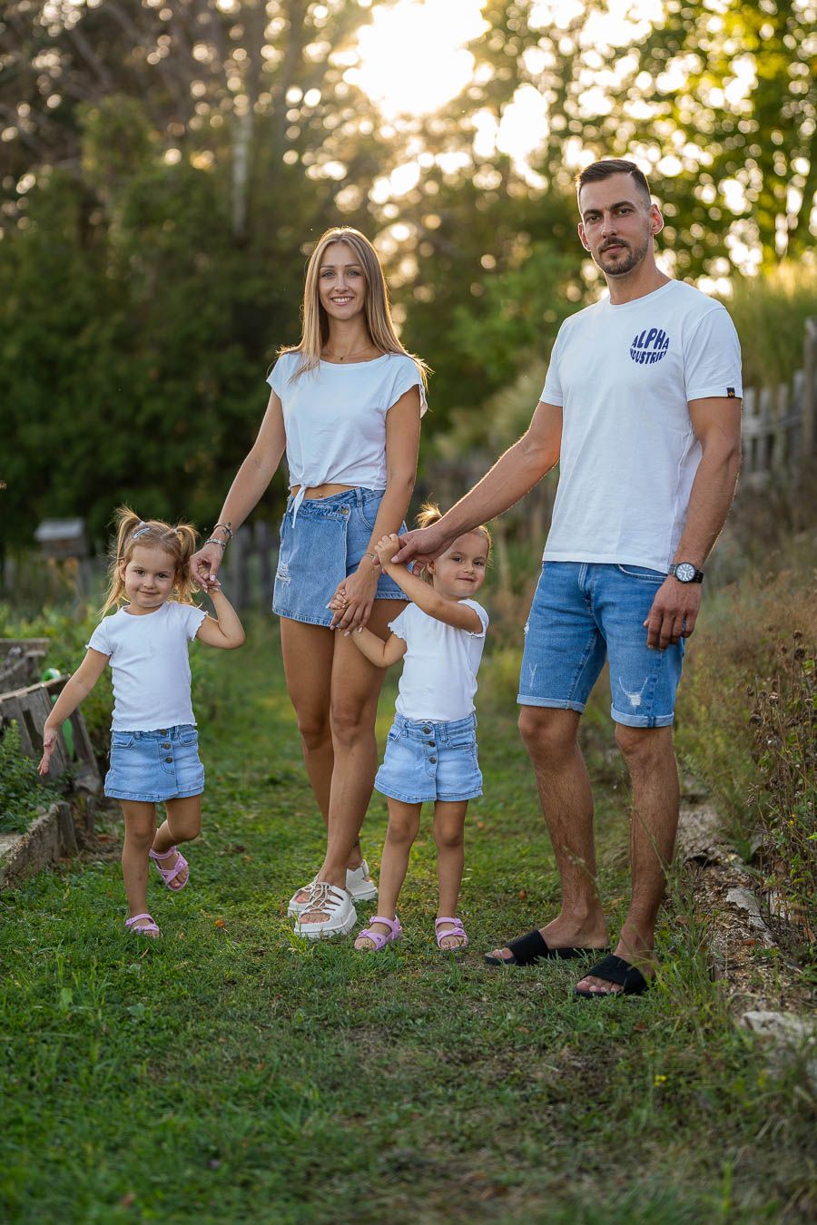 Happy family of four in matching white tops and denim shorts holding hands outdoors in a sunny green garden.