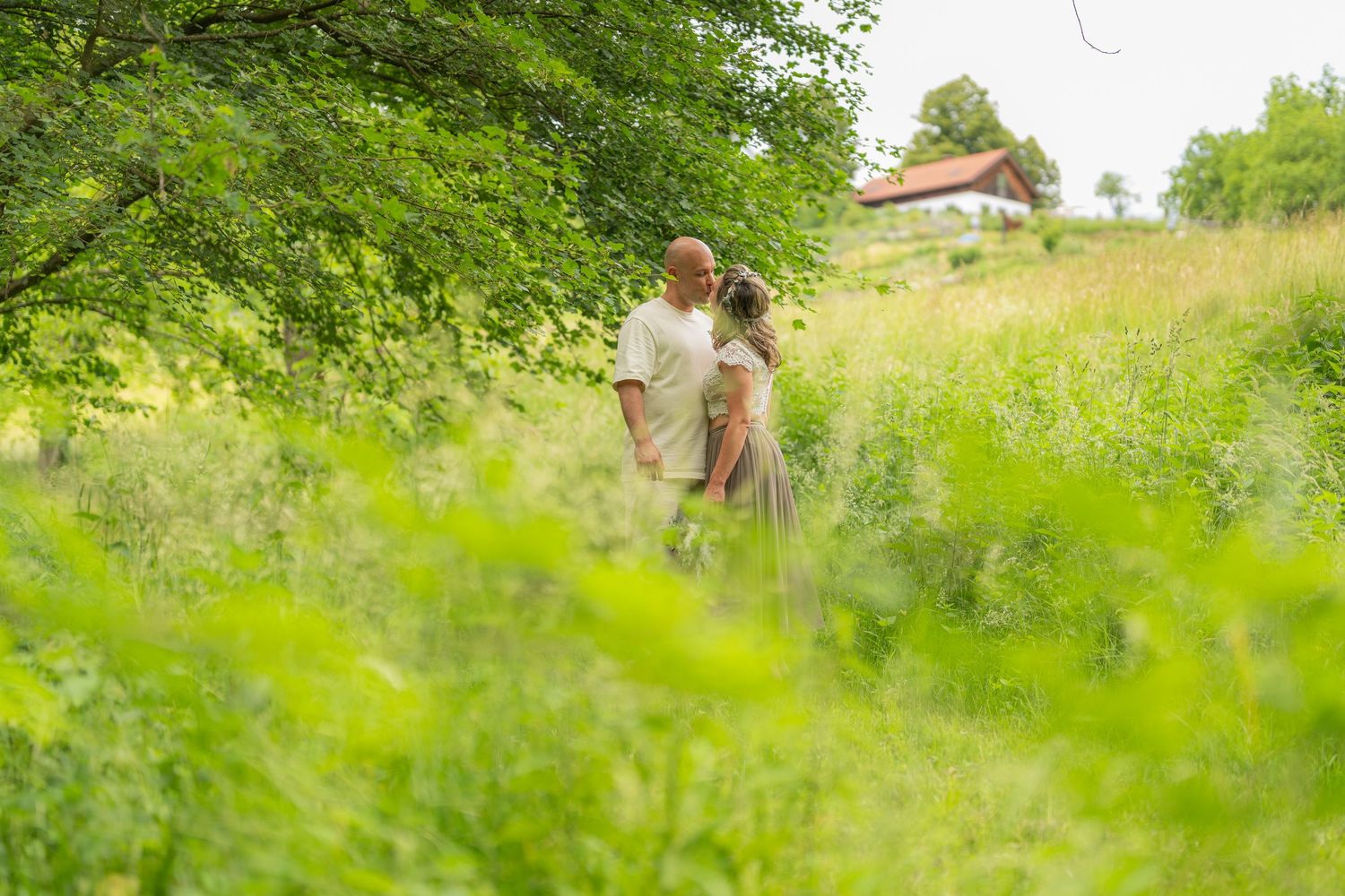 Couple embracing romantically in a lush green meadow surrounded by tall grass and trees on a sunny day.
