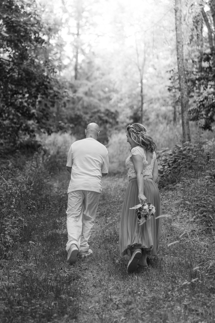 Black and white photo of a couple walking hand in hand down a forest path, bride holding a bouquet.