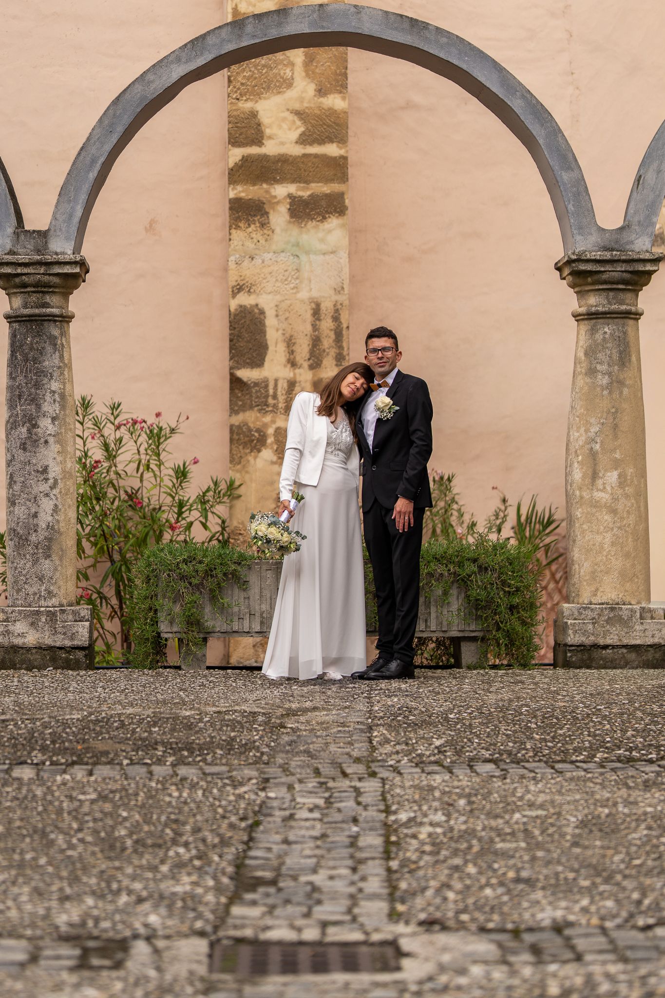 Wedding portrait under stone archways against textured wall and cobblestone ground.