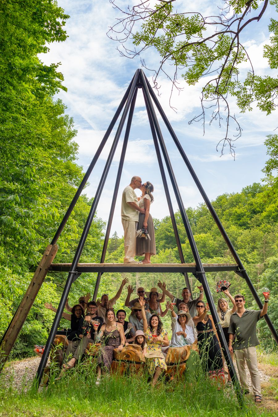 Couple kissing atop a metal pyramid sculpture surrounded by cheering group of friends in a lush green forest setting.