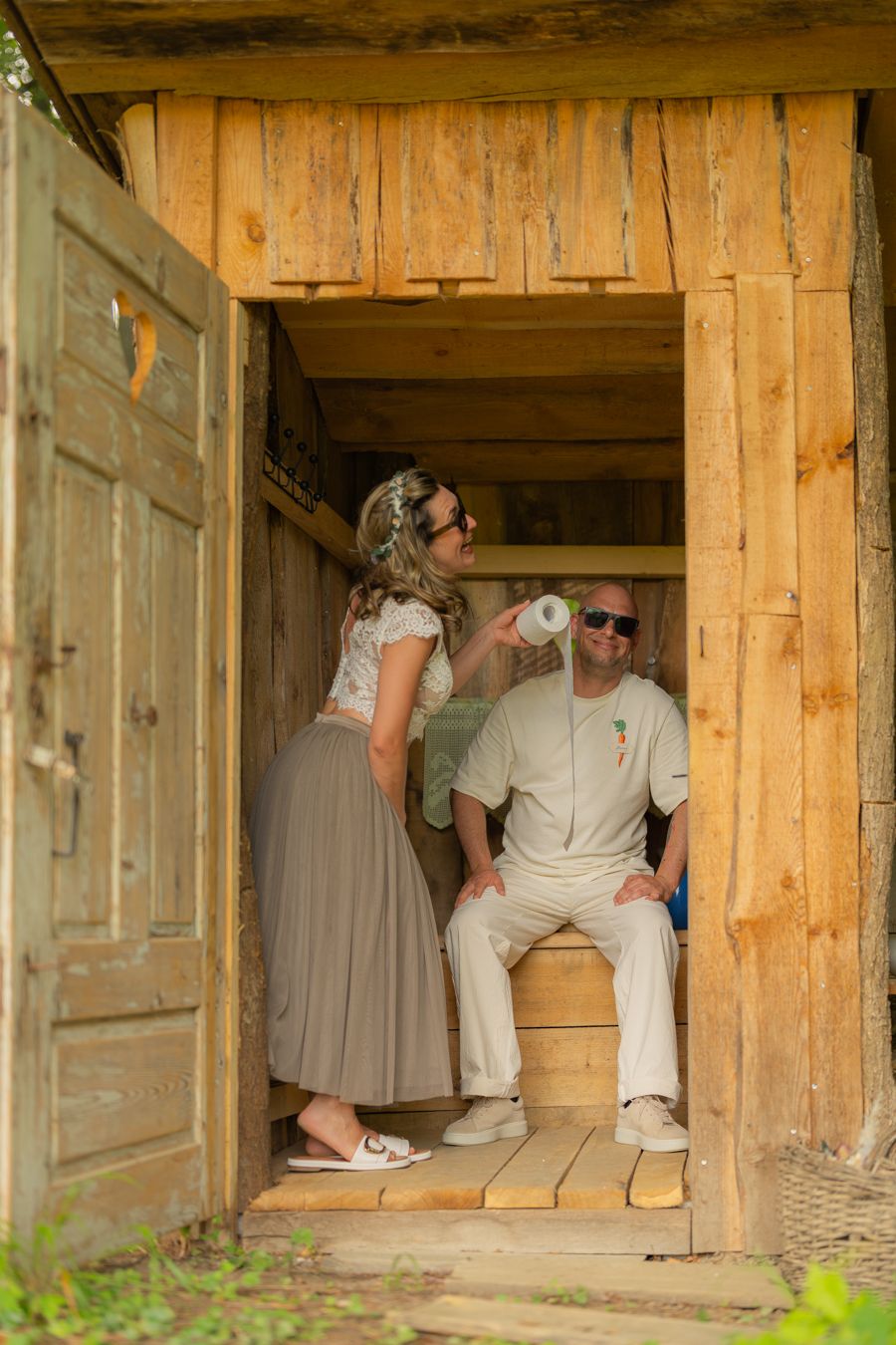 Couple relaxing in rustic wooden cabin doorway, woman offering drink to smiling seated man outdoors.