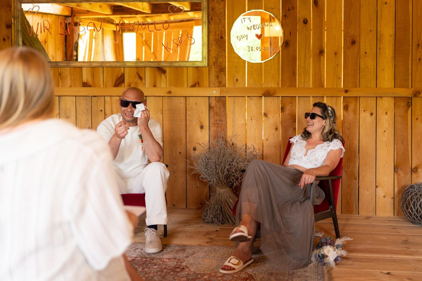 Two people wearing sunglasses relax in a rustic wooden cabin interior during a sunny day.